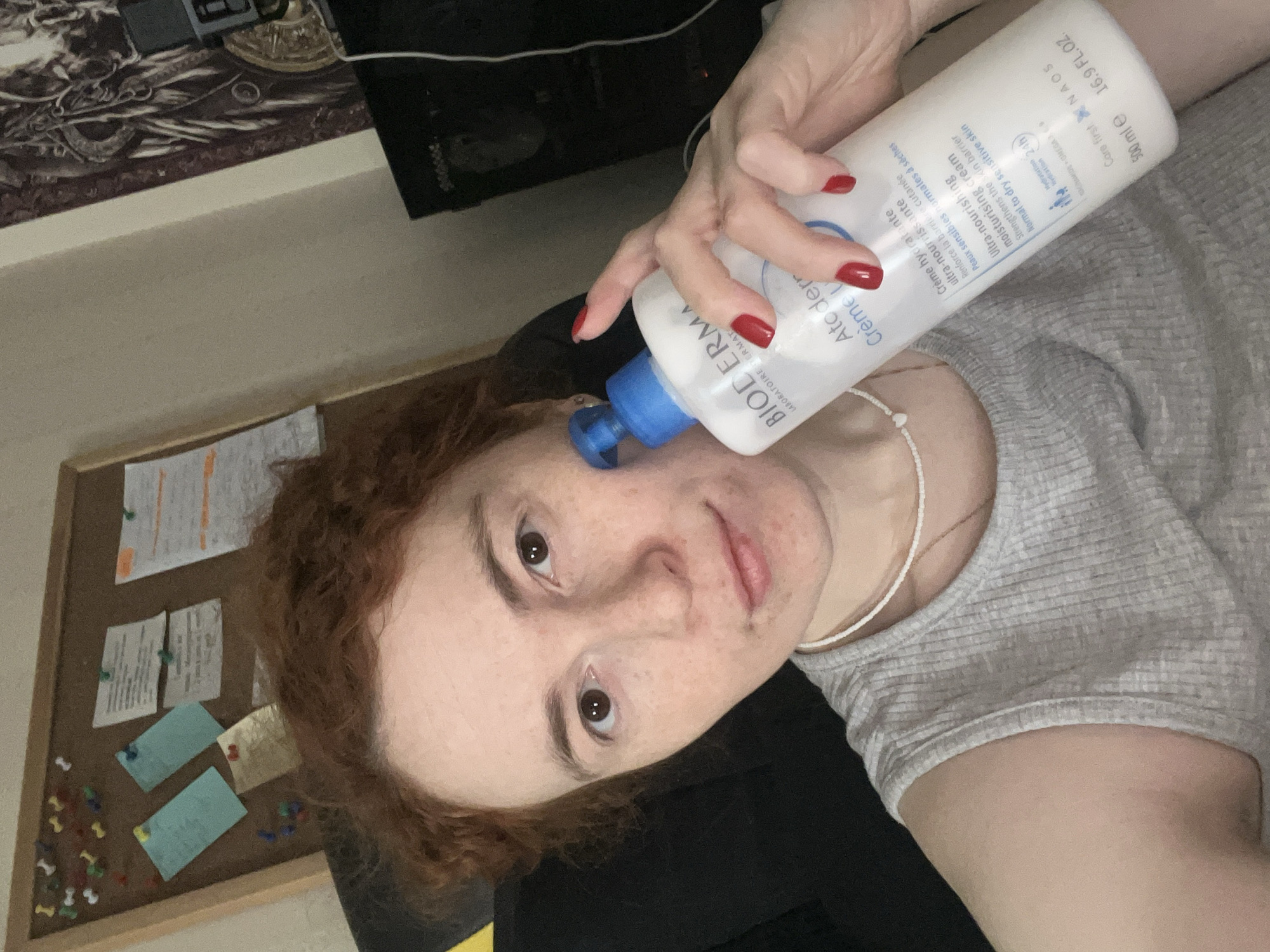 A fair-skinned woman with auburn, curly hair is shown in a selfie-style, angled shot. Her face, freckled and exhibiting clear skin, is the focal point as she looks directly at the camera. She is applying a bottle of BIODERMA Atoderm Creme to her face. Her fingernails are painted red. The background includes a corkboard with pinned notes or papers, a desk with items on it, and a gray t-shirt. The location is not determinable as Chaikovsky, Russia.