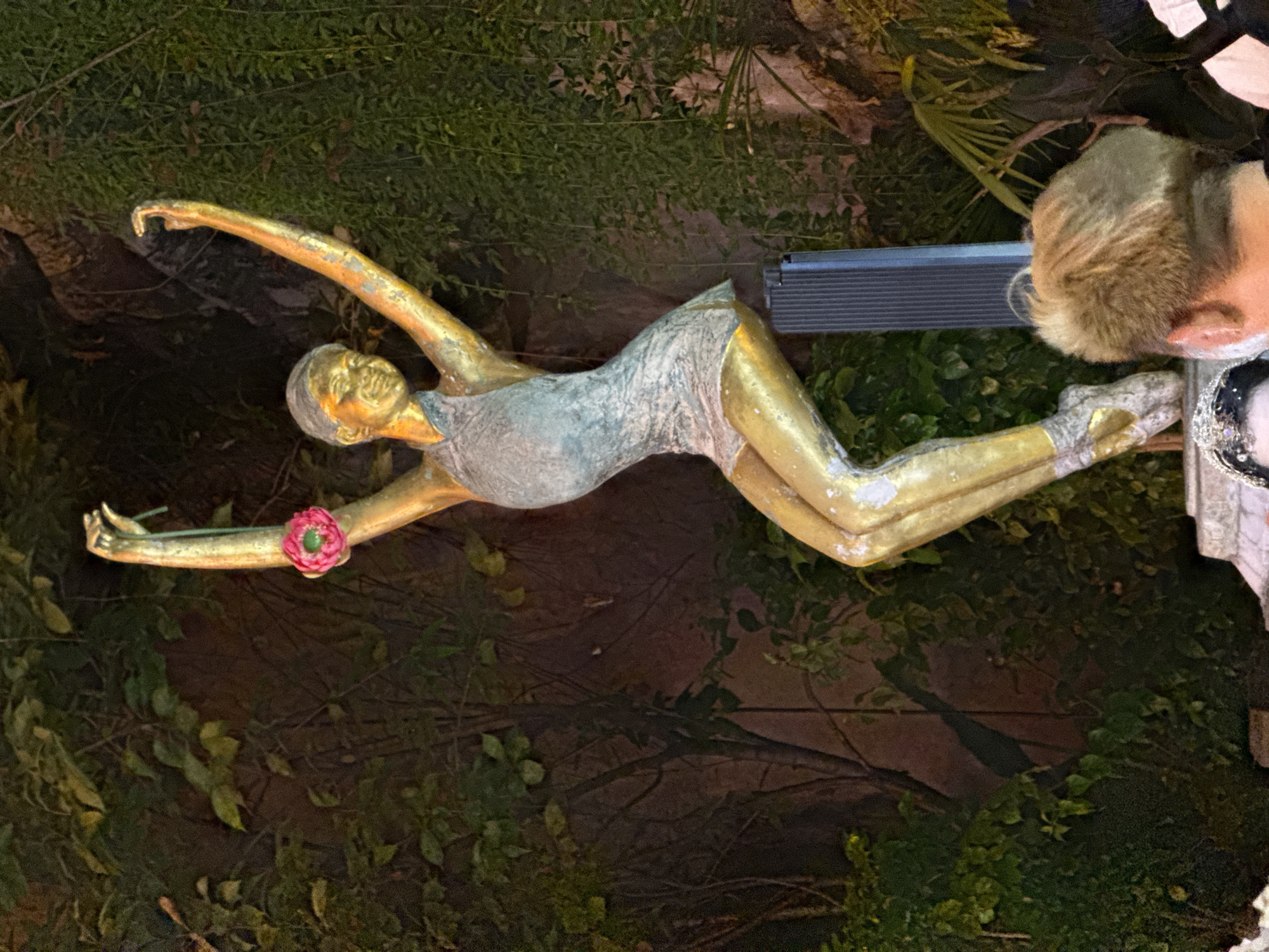 This is a medium shot photograph of a golden statue of a ballet dancer posed in a graceful, dynamic stance. The statue is in an outdoor setting, likely at night or in low light conditions, with lush green foliage and trees forming the background. The dancer statue is wearing a light-colored, short dress and ballet slippers. One arm is extended upwards, holding a delicate pink flower with a green stem. The other arm is also raised, bent at the elbow, with the hand open. The statue's face is smiling. In the foreground, the back of a person's head with blonde hair is visible, along with another person wearing a bejeweled headdress. A tall, dark, ribbed structure is partially visible behind the statue's legs. The overall scene suggests an artistic installation or monument in a public space within the Yuzhong District of China, possibly a park or plaza.