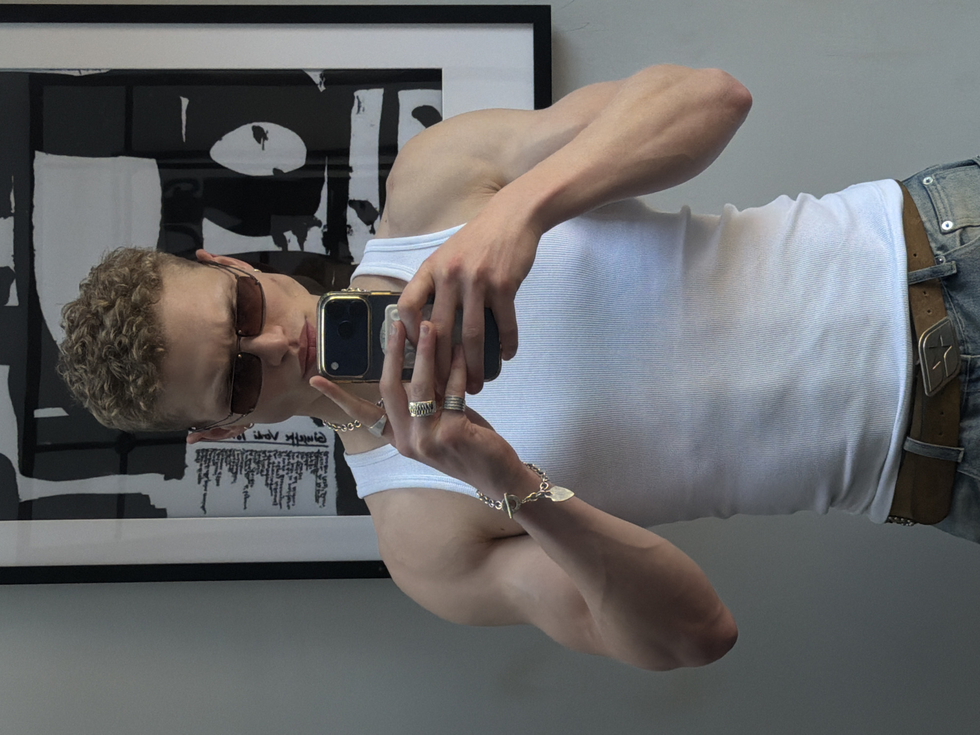 A man with curly blonde hair and sunglasses is taking a selfie in a room. He is wearing a white tank top and light blue jeans with a brown leather belt with a star-shaped buckle. He has multiple rings on his fingers and a silver bracelet on his wrist. He is holding a black smartphone with a gold case. Behind him is a black and white framed piece of art. The room is painted in a neutral color. The location is unknown and there is no evidence to suggest it is in London.