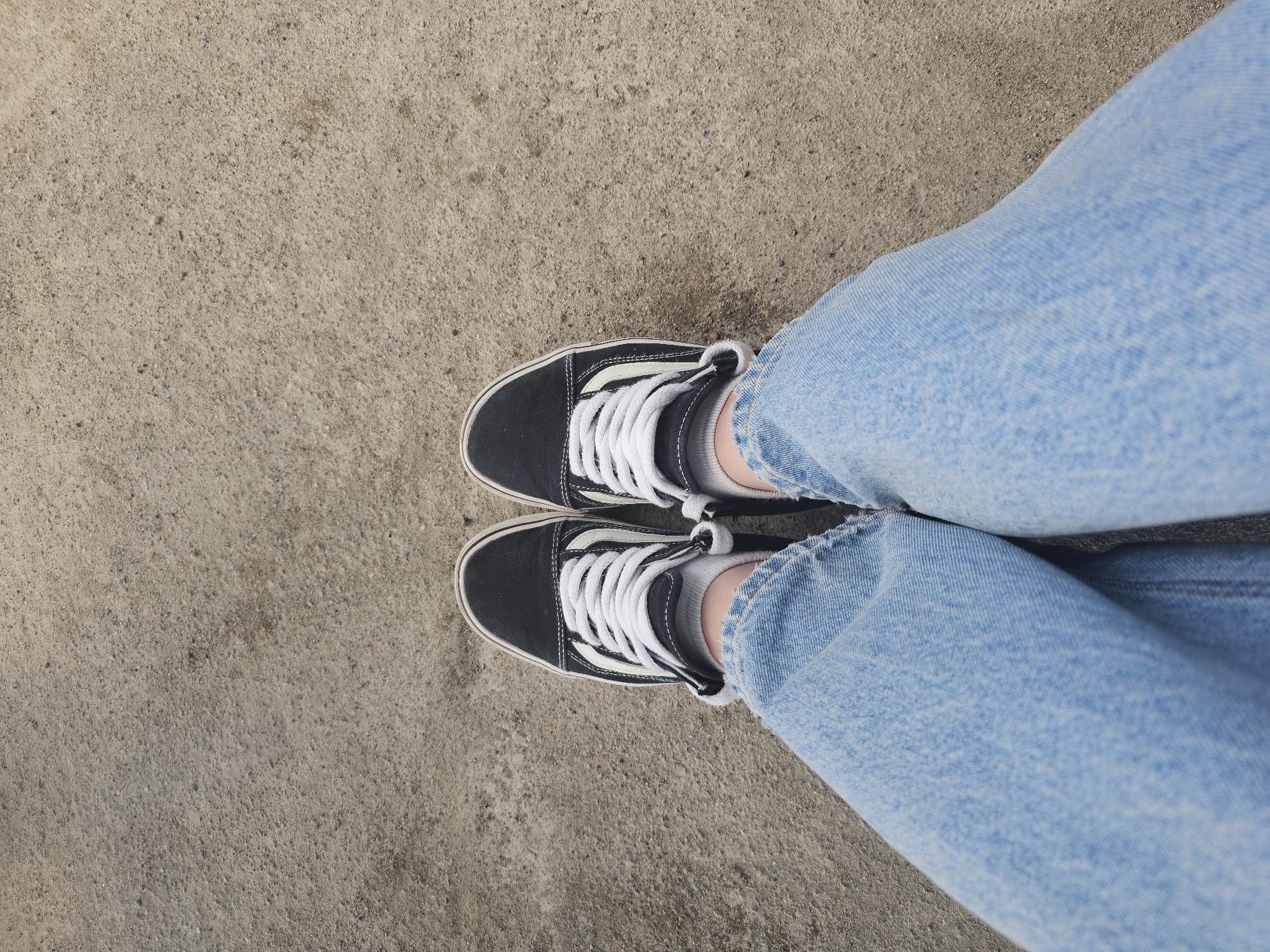 A top-down, eye-level shot shows a pair of feet wearing black Vans Old Skool sneakers with white laces and white socks, standing on a concrete surface. The jeans are light blue denim. The concrete is textured with small pebbles and appears to be an outdoor surface. The image was taken in Monchegorsk, Russia.