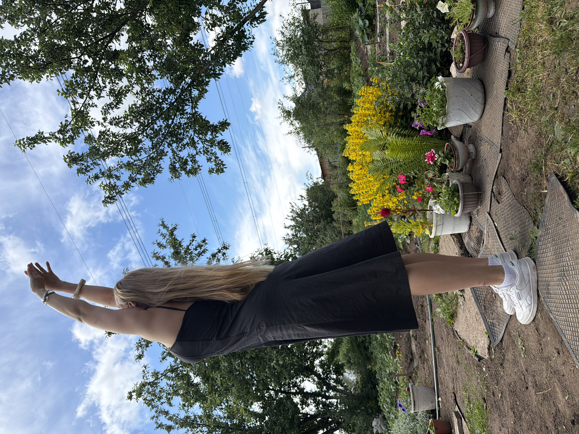 A young woman with long blonde hair is performing a yoga pose with her arms extended upwards in a garden located in Chaikovsky, Russia. She is wearing a black dress and white sneakers. The garden is lush with various plants, including yellow flowers and potted plants. The background shows trees, houses, and a cloudy blue sky with power lines.