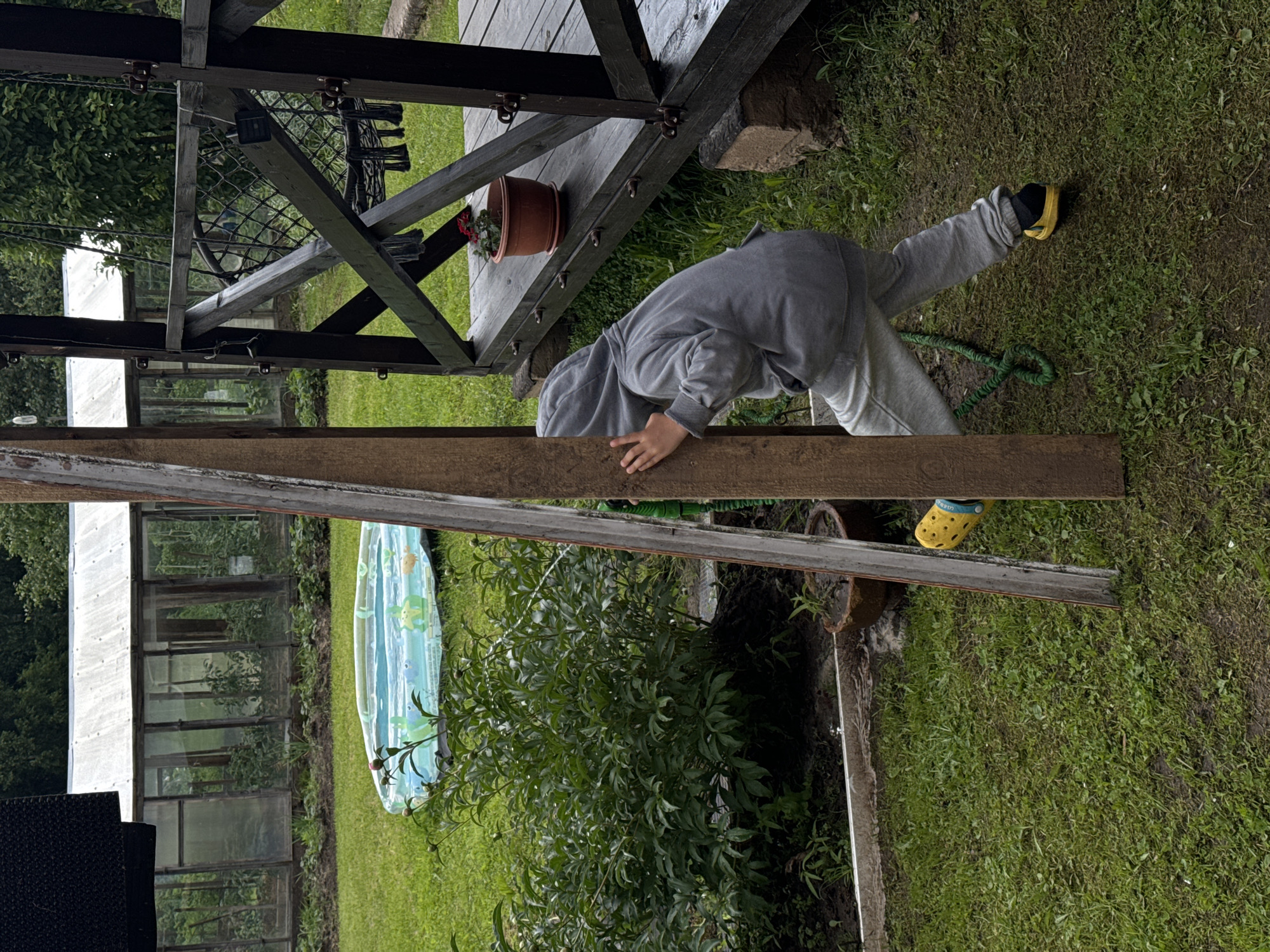 A child in a gray hooded sweatshirt and sweatpants, wearing yellow Crocs with black socks, is peeking around a wooden post in a grassy yard. The child's right hand is on the post, and their face is obscured by the hood. In the background, there is a greenhouse, a small inflatable pool with a cartoon pattern, a large green bush, a wooden porch with a swing, and trees. The scene appears to be in the city of Ponazyrevo, Russia.