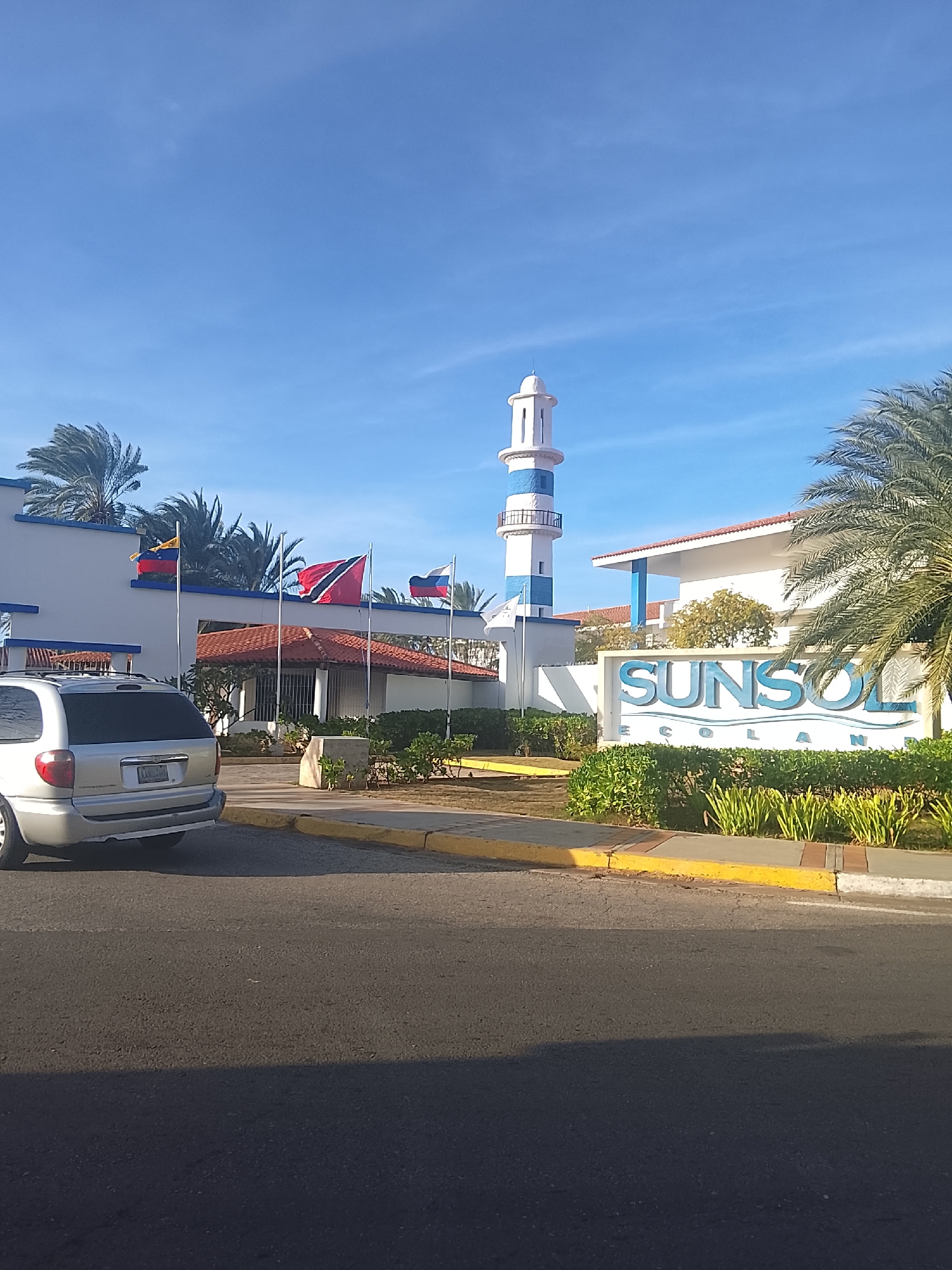 The image shows the entrance to the Sunsol Ecoland hotel in Altagracia, Venezuela. The hotel's sign, "SUNSOL ECOLAND," is prominently displayed. A tall, striped lighthouse structure stands behind the sign. Several flags, including those of Venezuela and Trinidad and Tobago, are visible on flagpoles near the entrance. A silver minivan is parked on the street in front of the hotel. The environment appears to be a sunny, coastal area with palm trees and well-maintained landscaping.