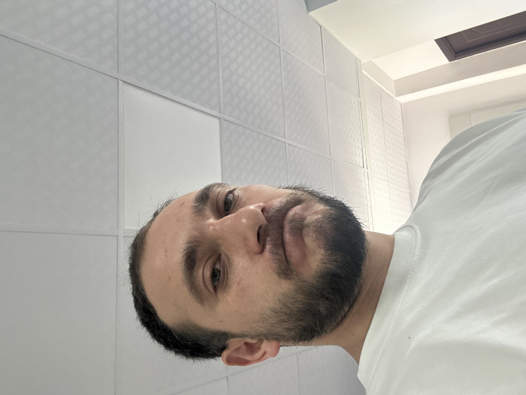 A man with dark hair and a full beard, wearing a white t-shirt, is depicted in an indoor setting. The image is taken from a low angle, showing his face angled slightly upwards and to the right. He has a neutral to slightly contemplative expression, looking towards the camera.
The background consists of light-colored, possibly white or light grey, wall panels. Some of these panels feature a subtle, repeating textured pattern, resembling small squares or diamonds. The panels are divided by vertical and horizontal lines, forming a grid-like structure. Above the man's head, a white ceiling is visible, with some architectural details including a recessed section and a dark brown rectangular vent or opening embedded within it.
No specific activity is taking place other than the man posing for the photograph. The lighting suggests an indoor daytime scene. There is no visible text in the image. The location context is Yerevan, Armenia.