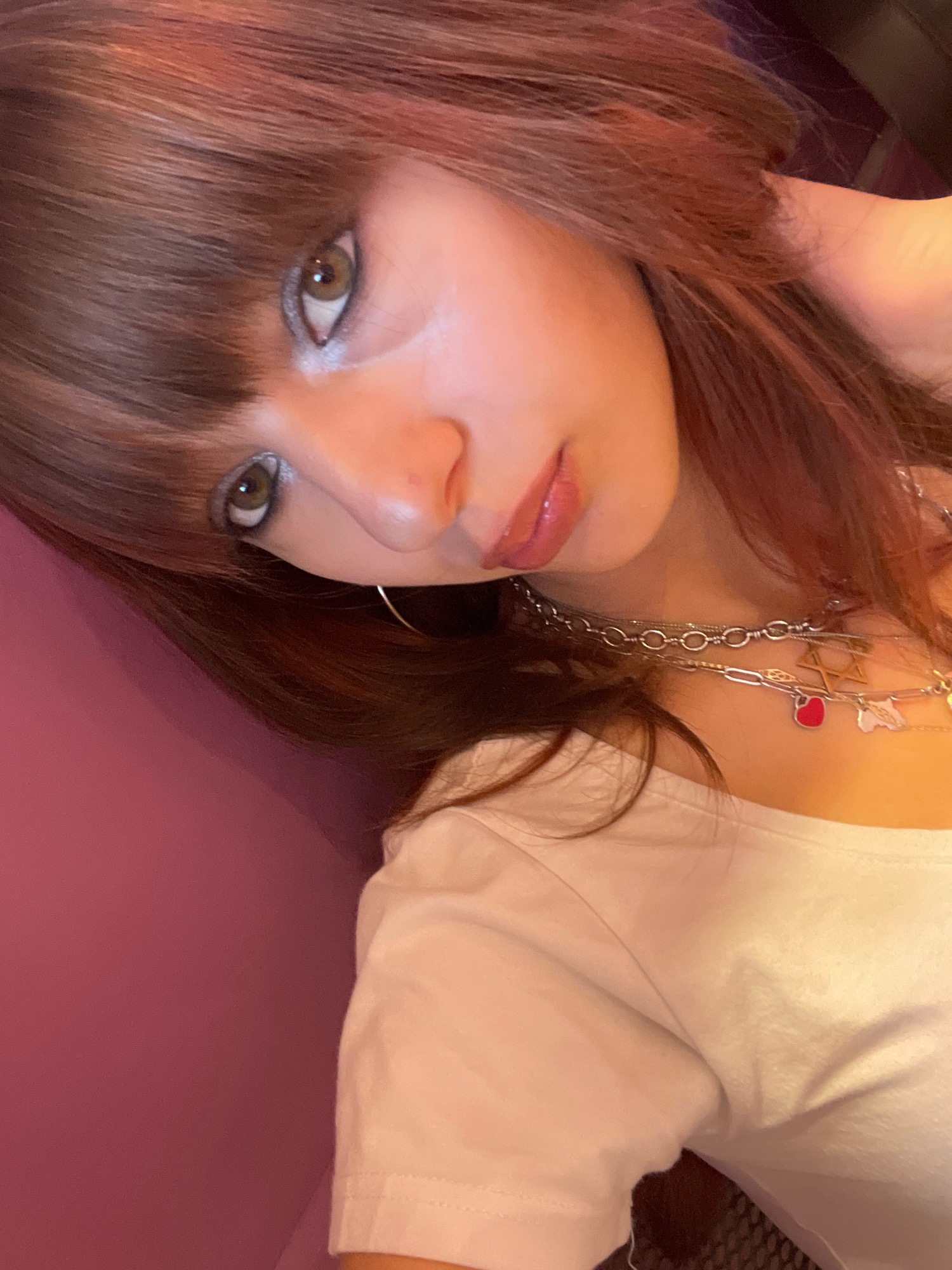 A young woman with shoulder-length, reddish-brown hair and bangs poses for a selfie, her head tilted to the side. She wears a white t-shirt and multiple silver necklaces, one featuring a Star of David charm and a red heart charm. Her makeup includes silver eyeshadow and lip gloss. The background is a textured pink surface. The image appears to be taken indoors. There is no discernible information within the image to suggest a specific location in Raanana, Israel.