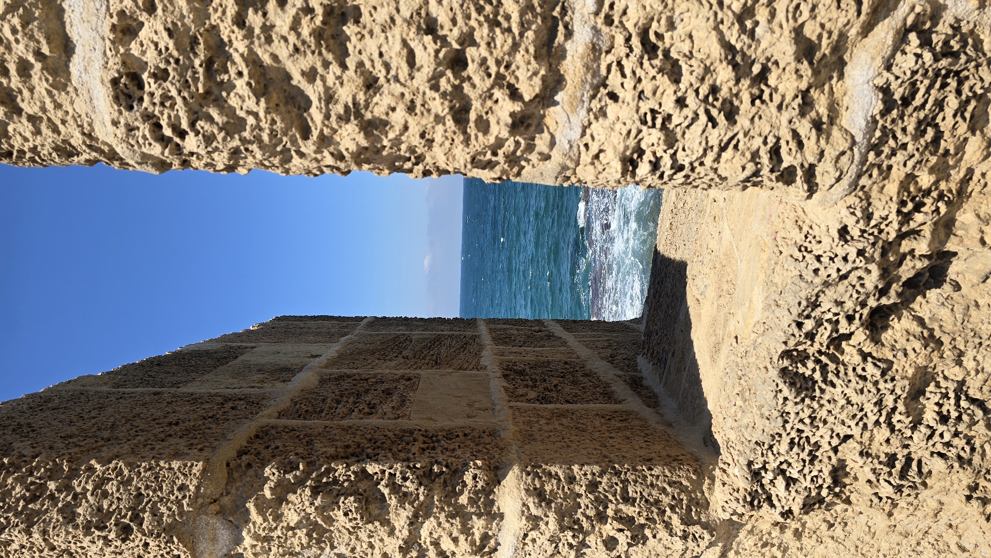 The image shows a view of the Mediterranean Sea through a gap in ancient stone walls, likely part of the Crusader fortifications in Acre, Israel. The walls are constructed from large, rough-hewn blocks of yellowish-brown stone, weathered and porous. The left wall is angled, revealing a section of its surface composed of smaller, rectangular blocks arranged in a grid pattern. The right wall is a vertical, textured column of the same rough stone. Between the walls, the sea is a vibrant turquoise blue, with white waves crashing against a rocky shoreline. The sky above is a clear, deep blue. The foreground consists of the sandy, uneven ground within the walls. There are no people or animals visible in the frame. The scene evokes a sense of history and the raw beauty of the coastal landscape.