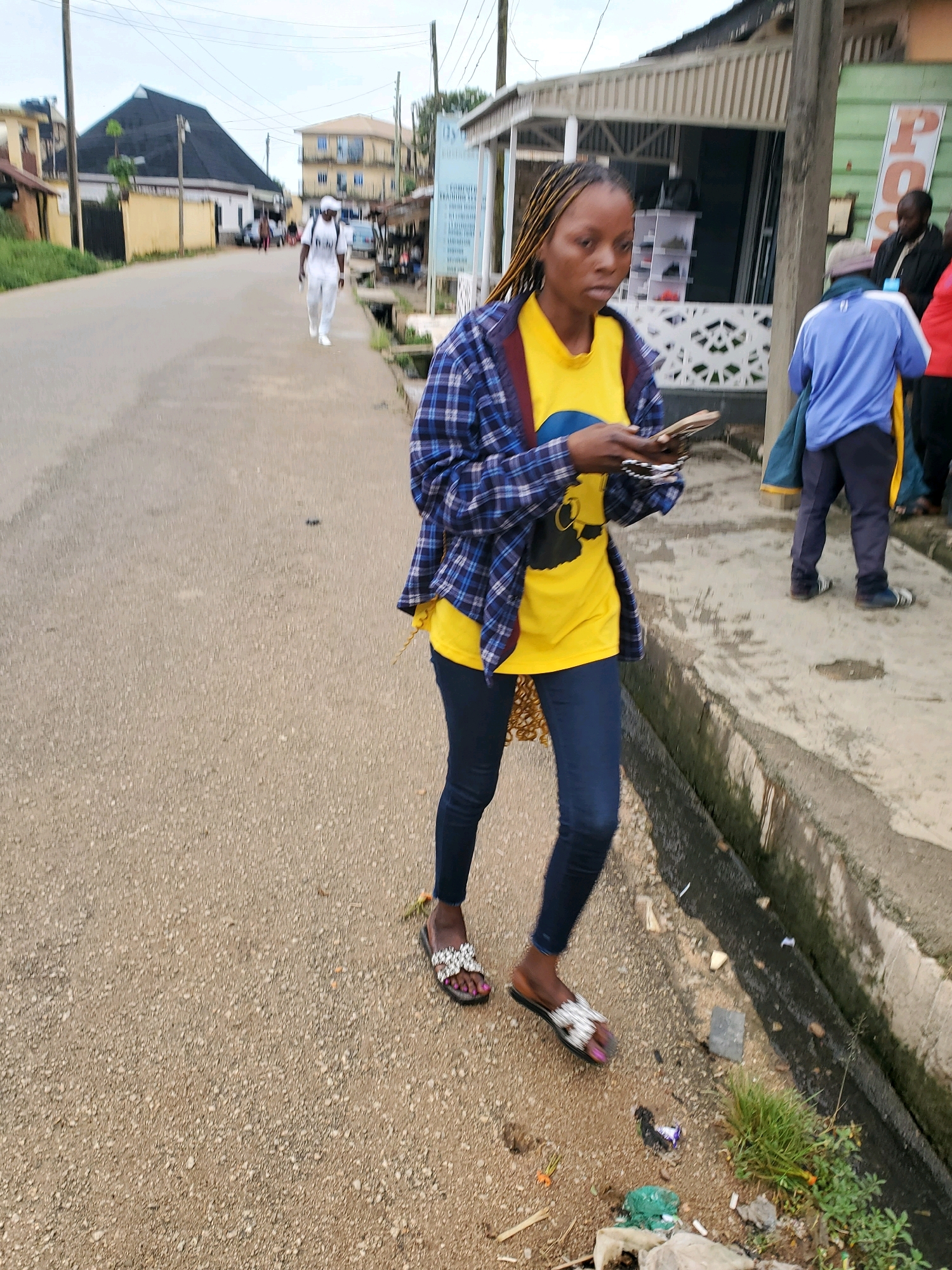 A young Black woman with braided hair walks on a paved street in Jos, Nigeria. She is wearing a yellow t-shirt, a blue and white plaid shirt, and dark blue jeans, with decorative sandals on her feet. She is holding a mobile phone and what appears to be cash in her hands. To her right, a partially visible Black man in a blue jacket walks away from the camera. Further down the street, another Black man in white clothing walks towards the camera, and a man in a blue shirt and dark pants walks past a building with shops. The street is lined with buildings, including a two-story building with balconies, and an overcast sky is visible. Debris is scattered along the edge of the street, including plastic bags, grass, and cigarette butts.