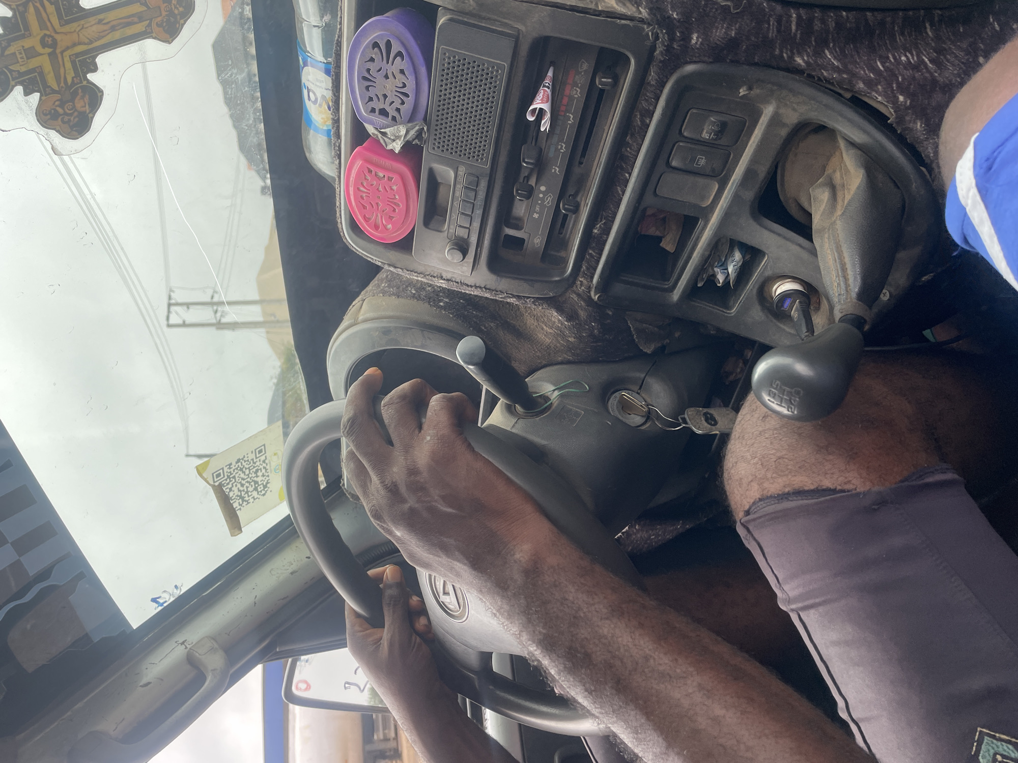 The image depicts the interior of a vehicle, likely a truck or van, as seen from the perspective of a passenger. A person's hands are visible on the steering wheel, indicating they are the driver. The dashboard is dusty and cluttered, with several items including air fresheners (pink and purple), a radio, and climate control knobs. A key is inserted into the ignition. To the right of the steering wheel, the gear shifter is visible, with markings indicating different gears. In the foreground, the driver's legs are visible, wearing grey shorts. The windshield shows a cloudy sky and some power lines and buildings in the distance. A QR code is affixed to the windshield near the top right. A religious sticker, a crucifix, is also present on the windshield. The scene appears to be within the city of Ogbunike, Nigeria, given the provided context.