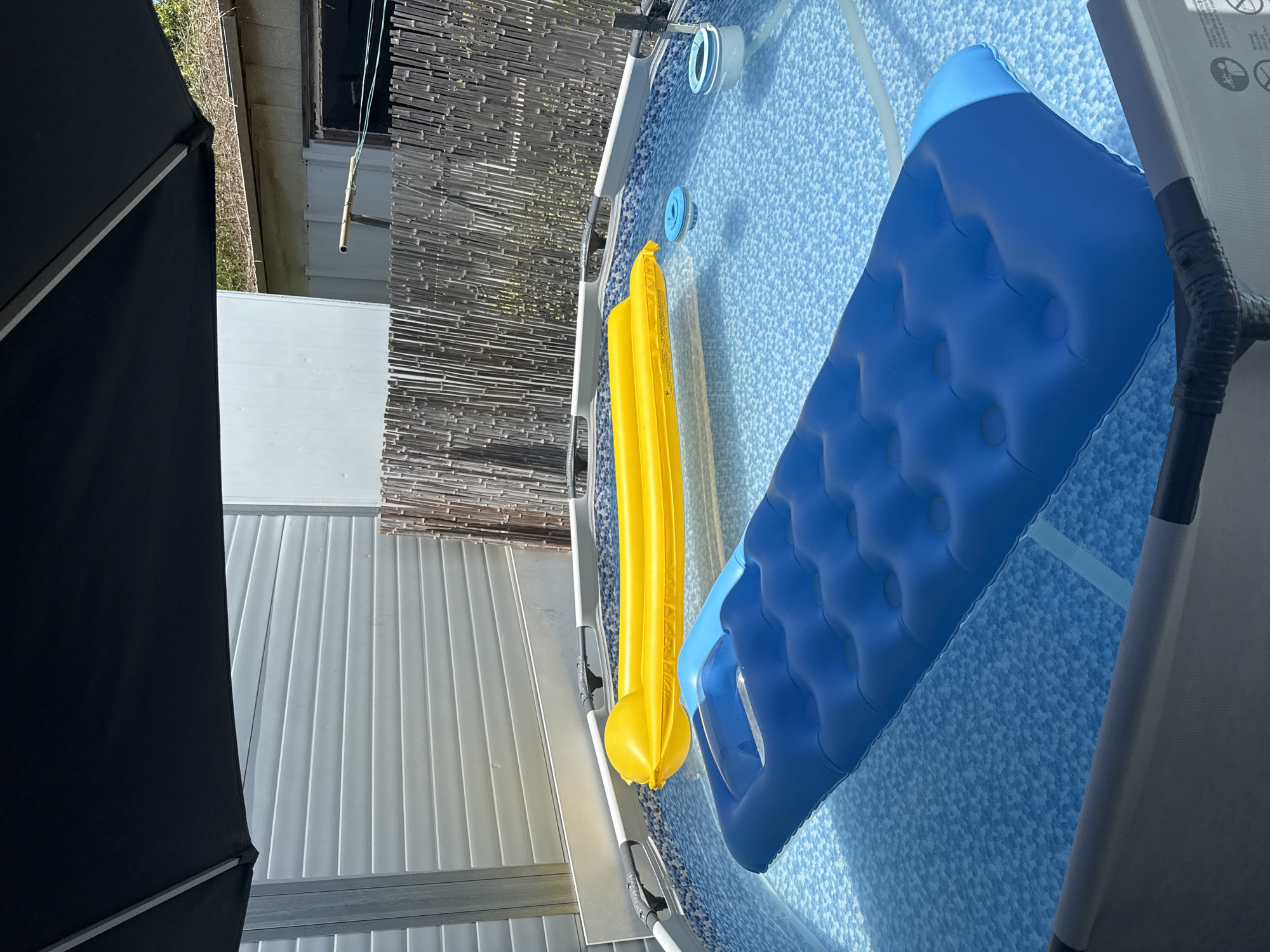 In Kiryat Gat, Israel, a private above-ground swimming pool is depicted on a sunny day, partially shaded by a large black umbrella. The pool is filled with clear blue water with a mosaic pattern at the bottom. Two inflatable pool floats are visible: a yellow one positioned in the background and a large blue one dominating the foreground. The pool is surrounded by a light-colored, textured fence and a grey siding structure. The general environment suggests a residential outdoor setting.