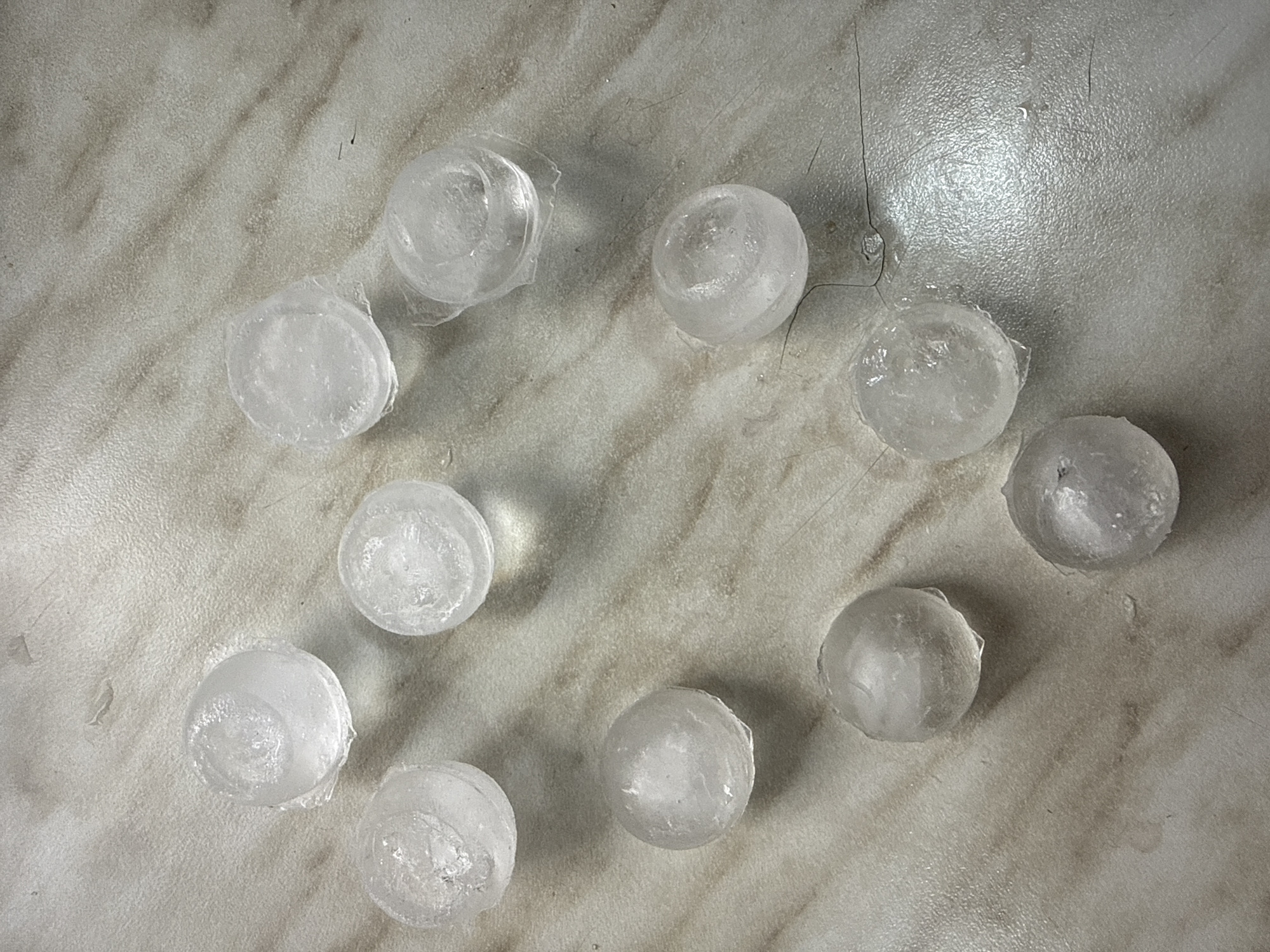 A top-down shot shows nine spherical ice cubes arranged in a heart-like shape on a marble countertop. The ice cubes are clear and some have small air bubbles. The countertop is light-colored with subtle grey veining.