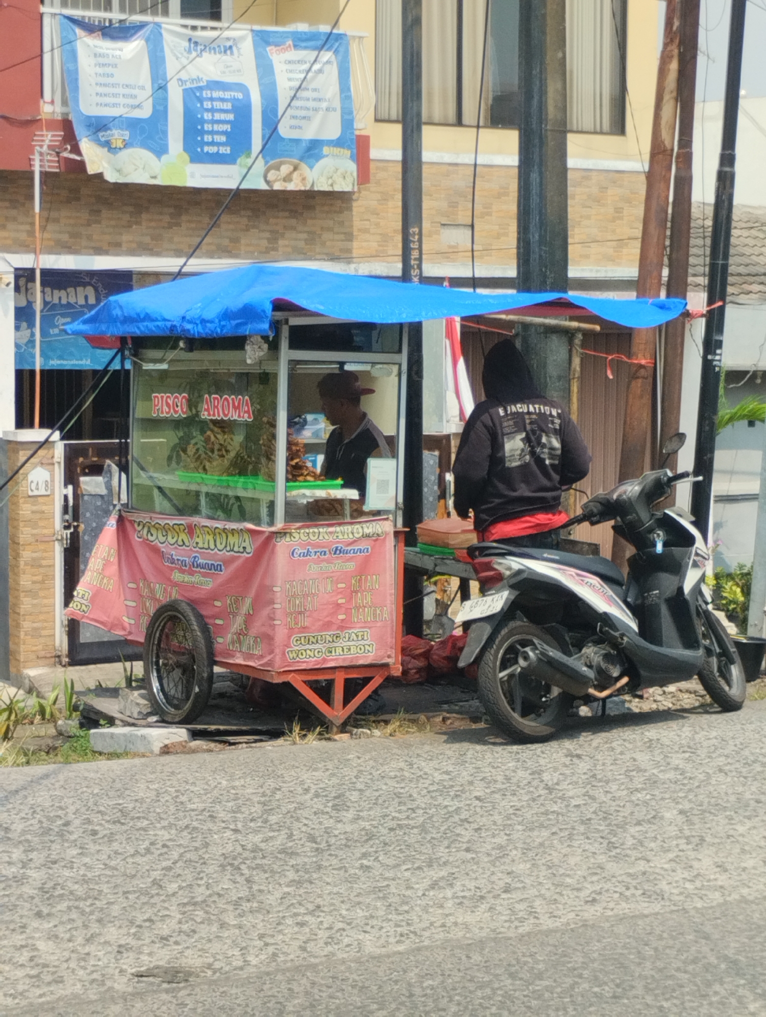 The image depicts an outdoor scene in front of a building, possibly in the city of Bekasi, Indonesia. A food cart selling "PISCO AROMA" is prominent in the foreground, with a blue tarp providing shade. The cart displays various food items and a red banner with text in Indonesian, listing ingredients like "KACANG IJO," "KETAN," "COKLAT," "TAPE," and "NANGKA," along with "GUNUNG JATI WONG CIREBON." A person wearing a dark hoodie with "EJACUATION" written on the back stands behind the cart. A motorcycle is parked to the right of the cart. The background shows a building with windows and a banner advertising food and drinks. The street in front of the cart appears to be paved.