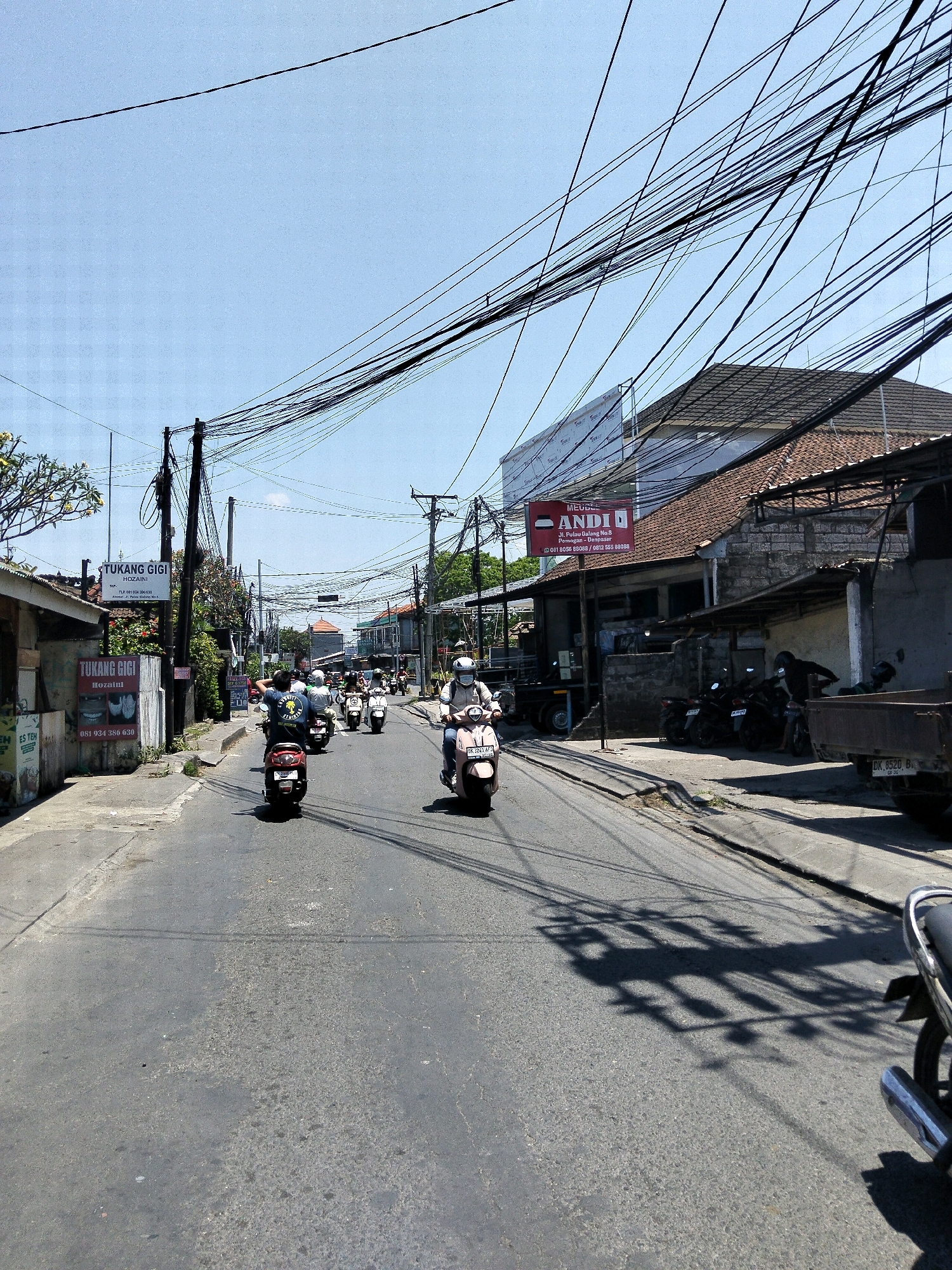 The image depicts a street scene in Denpasar, Indonesia, characterized by an abundance of overhead electrical wires creating a dense, tangled network against a clear blue sky. The street is paved with asphalt and appears to be a busy thoroughfare with moderate traffic. Several people are riding motorbikes, some of whom are wearing helmets.
On the left side of the street, there are buildings with signs, one of which reads "TUKANG GIGI HOZAINI," indicating a dentist's office. Another sign above it displays "TUKANG GIGI." Further down the street, another building has a red sign with "MEUBEL ANDI" and an address.
The right side of the street features more buildings, some with tiled roofs, and a parked truck. The environment suggests a commercial or mixed-use area within the city. The lighting indicates it is daytime, with sunlight casting shadows on the road. The overall impression is one of urban infrastructure and daily life in Denpasar.
