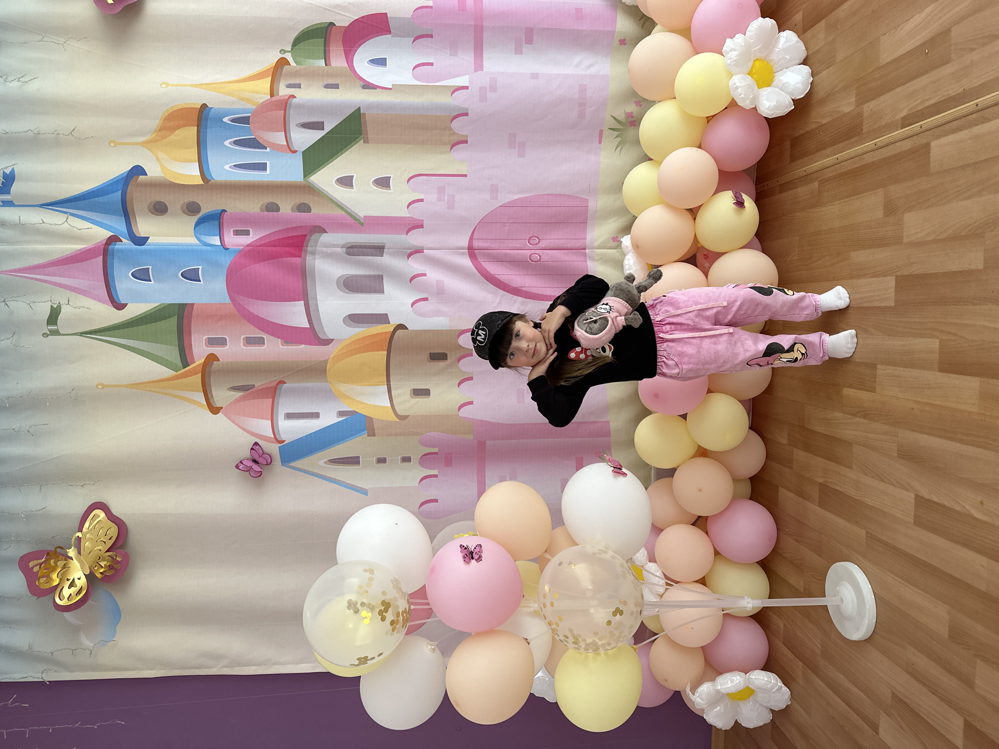 A child is lying on the floor in front of a backdrop of a cartoon castle, surrounded by balloons. The location is Chaikovsky, Russia.
The child is wearing a black long-sleeved shirt, pink patterned pants, white socks, and a black baseball cap with a white "M" logo. They are holding a stuffed animal.
The backdrop depicts a colorful cartoon castle with towers in various shades of pink, blue, yellow, and green.
Several balloons in pastel shades of pink, yellow, and white are arranged around the child and along the backdrop. Some are transparent and filled with gold confetti. A few decorative butterflies are attached to the backdrop and some of the balloons. There are also some large white flower-shaped balloons.
The floor is made of wood. A white balloon stand is visible.