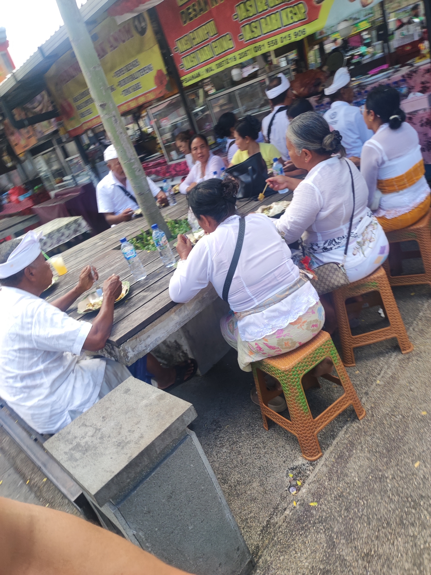 The image captures a group of people eating at an outdoor eatery in Gianyar, Indonesia. They are seated at a long wooden table, some on stools, and are dressed primarily in white traditional clothing, including head coverings for the men. Plates of food and water bottles are visible on the table. The background shows street-side vendors and signage in Indonesian. The location appears to be a bustling street with pedestrian traffic.