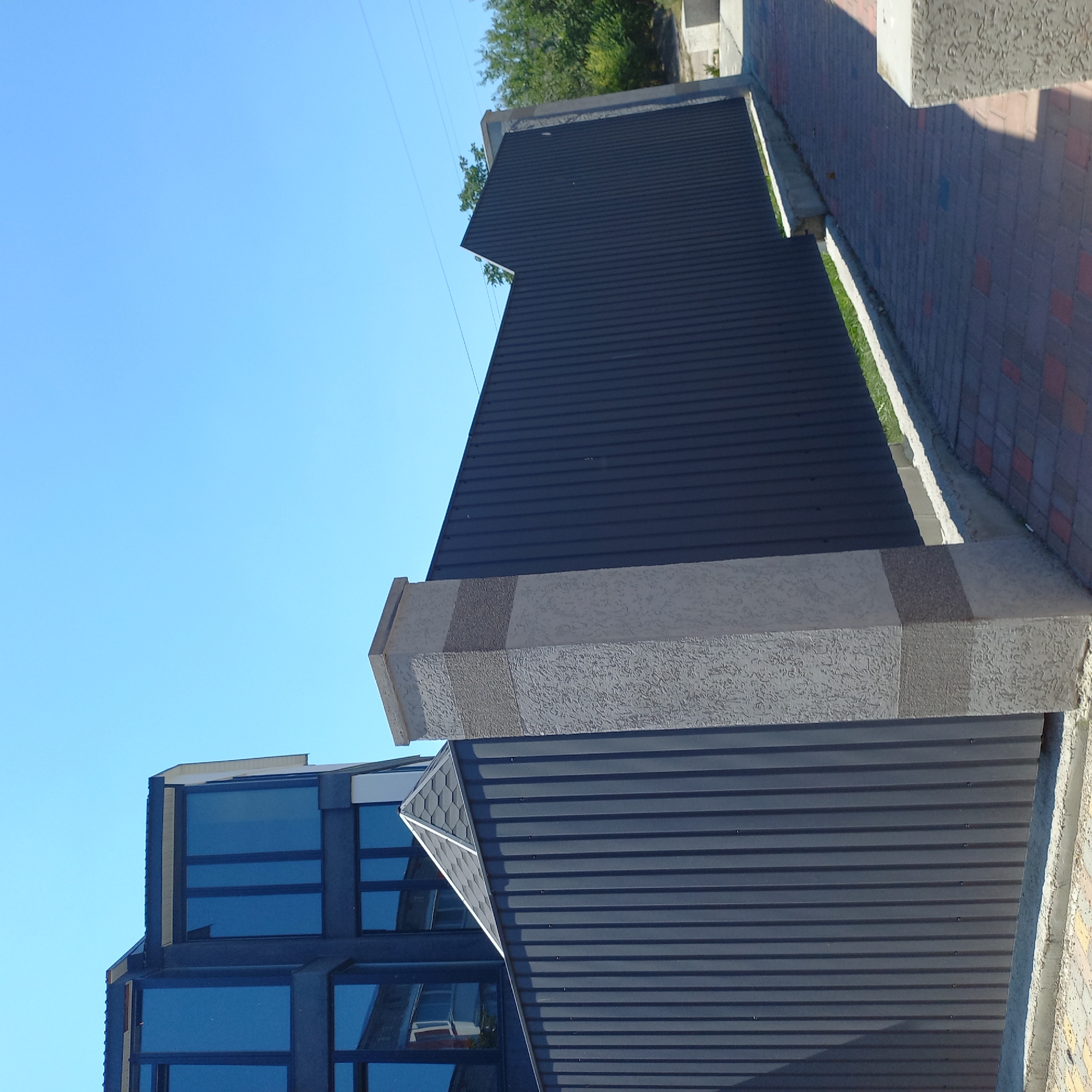 This image depicts a modern building with dark-framed, reflective windows situated behind a tall, dark gray corrugated metal fence. The fence is supported by concrete pillars, one of which is visible in the foreground, textured and banded with different shades of gray. The fence extends horizontally across the frame, with a peaked section on the right side. To the right of the fence, a paved walkway made of reddish-brown and gray rectangular pavers leads away from the viewer. Beyond the fence and building, there are trees and a clear blue sky. The scene appears to be a residential or commercial property under construction or recently completed, likely located in the city of Kropyvnytskyi, Ukraine, given the context.