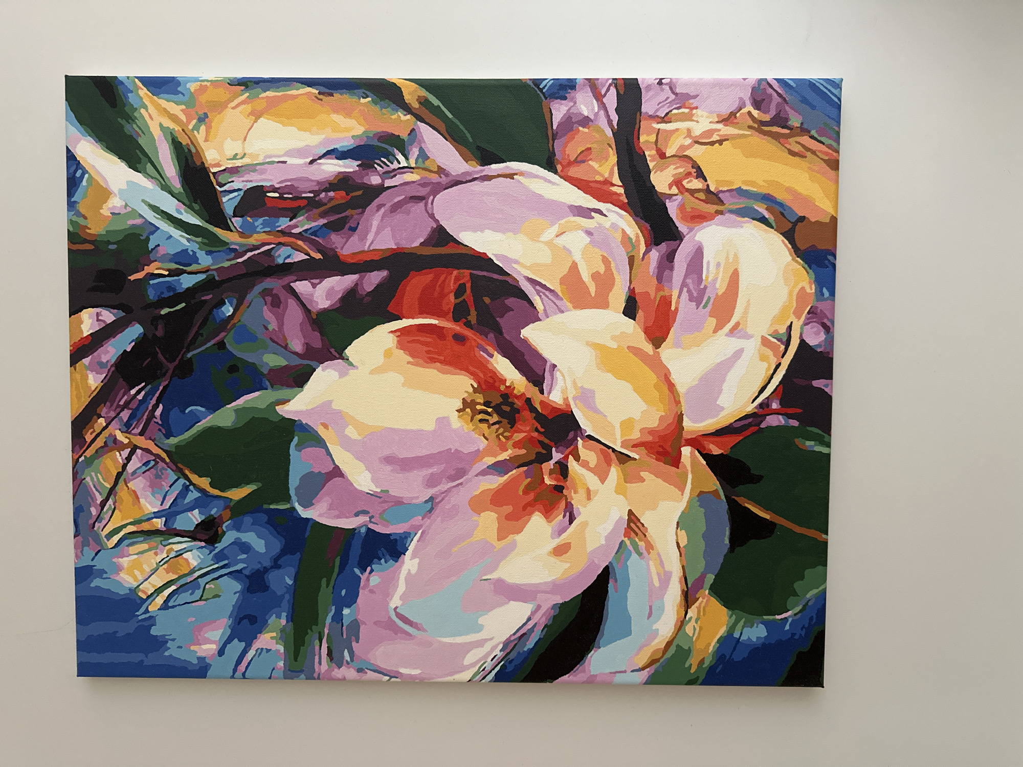 The image shows a painting of a magnolia flower with vibrant, stylized petals and surrounding foliage. The flower is rendered in shades of pink, yellow, orange, and white, with a darker center. The background is a mix of blues, purples, and greens, suggesting leaves and branches. The painting is displayed on a white wall. There is no visible indication of the city of Yessentuki, Russia.