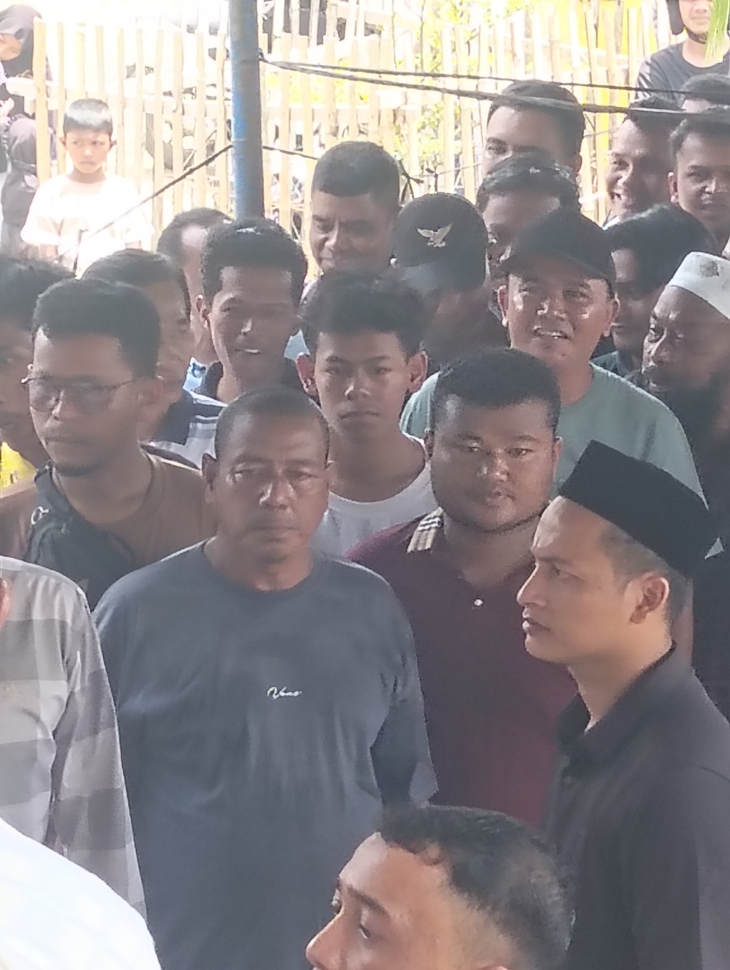 In the city of Grong-Grong, Indonesia, a group of approximately ten men and one boy are gathered outdoors, possibly at a community event or gathering. The men are of various ages and ethnicities, and most are wearing casual clothing. Some are looking directly at the camera, while others are looking in different directions. The background consists of bamboo fencing and possibly some structures. The lighting suggests it is daytime.