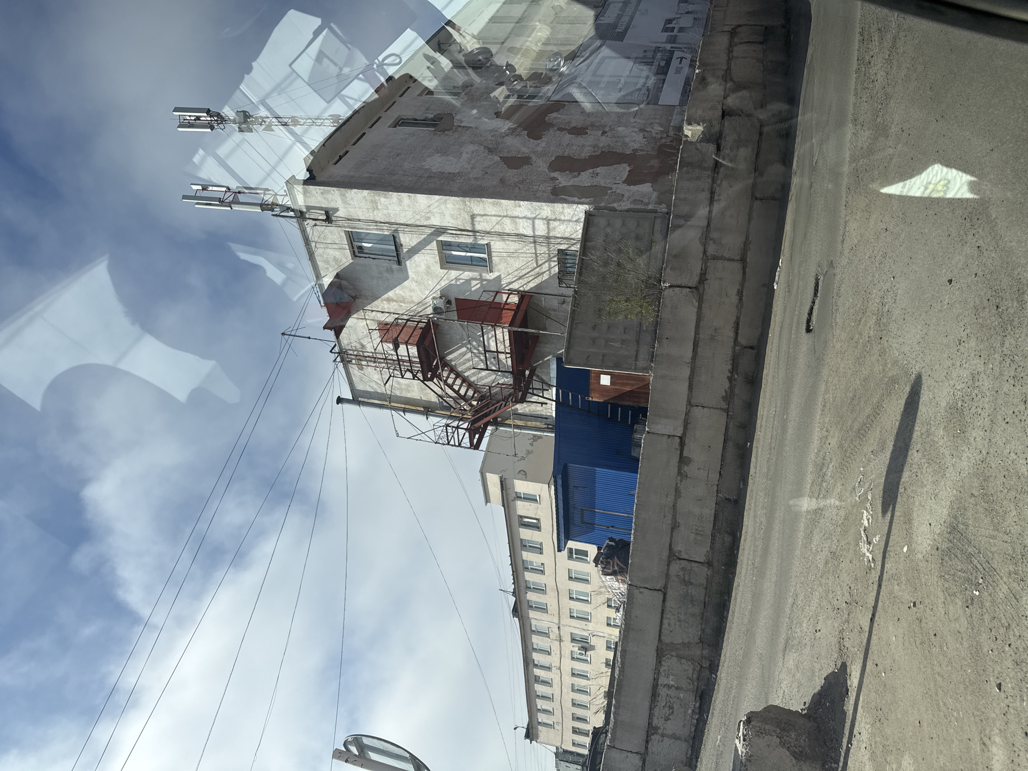 This image depicts an urban outdoor scene in the city of Magadan, Russia, captured from the interior of a vehicle, evidenced by prominent reflections on glass. The environment is characterized by multiple buildings, an uneven ground surface, and a partly cloudy sky.
In the right half of the image, a three-story (at least) building with a light grey, worn concrete or stucco facade is visible. Its exterior shows significant signs of decay, with large, irregular reddish-brown patches of peeling material. Multiple rectangular windows with light-colored frames are present on its visible side. An external red metal fire escape or service staircase is affixed to this building, leading to red-painted doors on at least two levels. The roof of this building hosts two cellular communication tower structures, one taller and positioned slightly behind the other.
To the left of the prominent grey building, and partially obscured, is a longer, lower, two-story structure with a cream or beige facade and a uniform row of rectangular windows. This building appears to be in better condition than the one on the right. Between these two buildings, and slightly in front of the longer one, stands a bright blue, single-story utility building or shed, constructed of corrugated metal.
A substantial concrete block retaining wall runs across the mid-ground, separating the elevated area of the buildings from the foreground. The blocks are large, rough-textured, and grey.
The foreground consists of a degraded asphalt road surface mixed with dirt and gravel. Tire tracks are visible on the ground, along with small pieces of debris. A notable pothole is present in the asphalt. Shadows are cast on the ground, indicating direct sunlight from the right. A small, sparse green bush grows near the base of the concrete wall.
The sky above is light blue with scattered white cumulus clouds. Multiple thin black power lines or communication cables traverse the upper part of the frame.
Prominent reflections on the vehicle's window include distorted images resembling a human head and shoulders, geometric shapes indicative of vehicle interior elements, and faint, inverted text, including what appears to be a Cyrillic "Вход" (Entrance) and an arrow. No human figures or animals are directly visible in the scene.