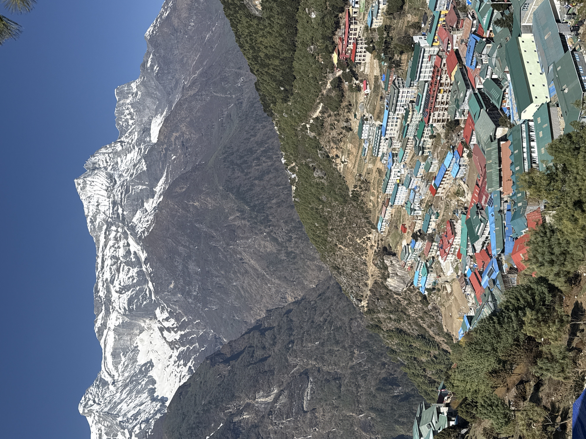 This image presents a stunning, wide-angle view of a vibrant mountain village, identified as Namche, Nepal, nestled dramatically in the Himalayan landscape. On the left side of the frame, a colossal, snow-capped mountain peak rises majestically, its jagged, grey, and white rock faces sharply defined against a brilliant, clear blue sky. The lower slopes of this mountain are rugged and dark.
To the right, the village spreads across a steep, forested hillside. It features a dense cluster of buildings with distinctively colorful roofs in shades of red, blue, green, and white, creating a striking contrast with the natural surroundings. Many structures appear to be multi-story. Scattered evergreen trees dot the hillsides, particularly around the village and in the foreground. The terrain is rocky and challenging, characteristic of high-altitude regions.
The scene is illuminated by bright daylight, suggesting a sunny morning or early afternoon with no clouds. No specific activities or people are discernible, presenting a static, grand overview of the settlement. The image conveys the scale of the mountains and the intricate way human life adapts to such an imposing natural environment. There is no visible text in the media.