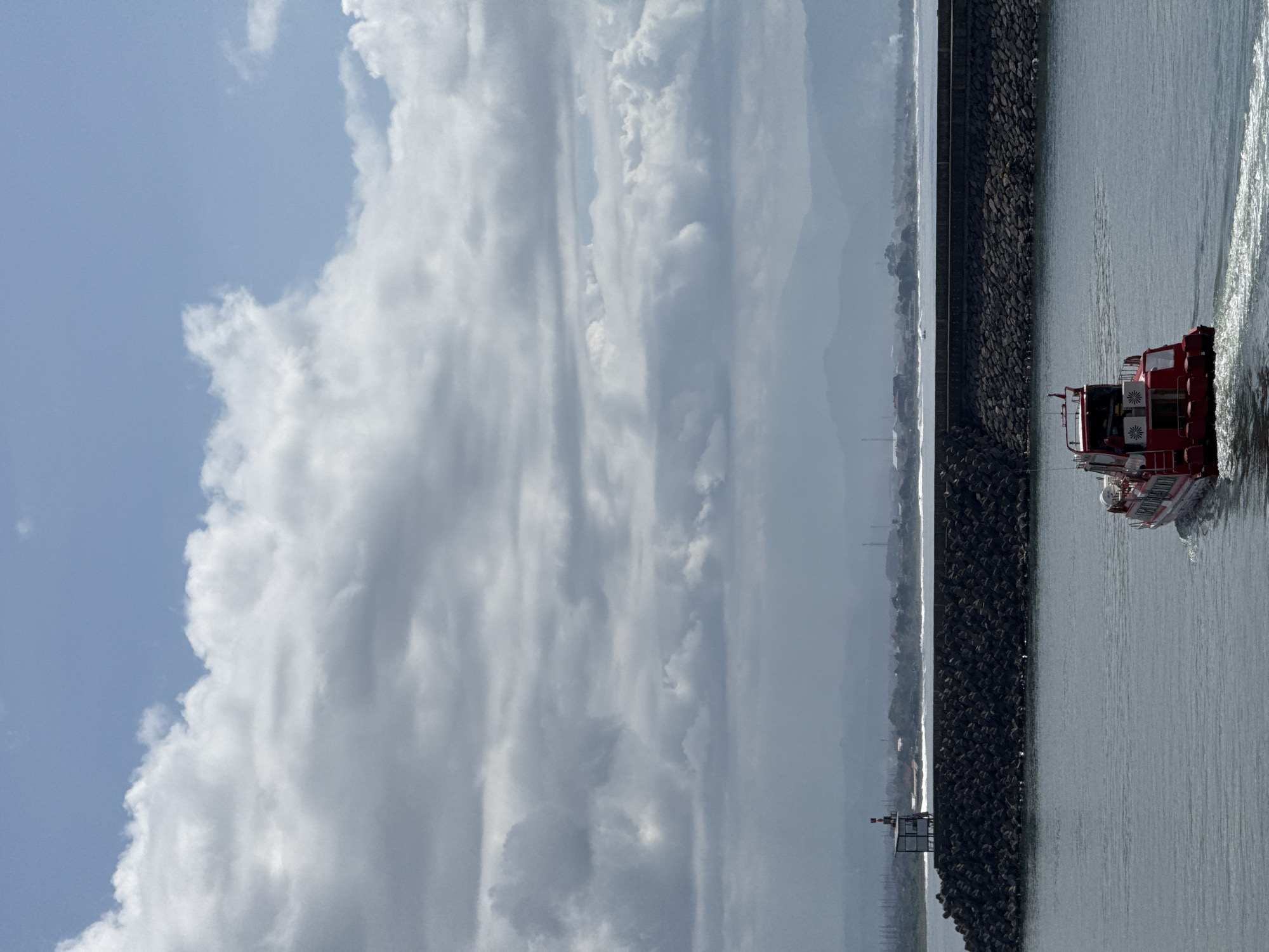 The image captures a red and white rescue boat navigating a channel in Denpasar, Indonesia. The boat is moving through the water, leaving a wake behind it. The channel is lined with a dark grey rock seawall, and beyond that, a hazy coastline is visible, hinting at a distant landmass. The sky is dominated by large, white cumulus clouds, contrasting with patches of blue sky. A small structure, possibly a navigational aid, is seen near the seawall. The lighting suggests a bright, possibly sunny day.