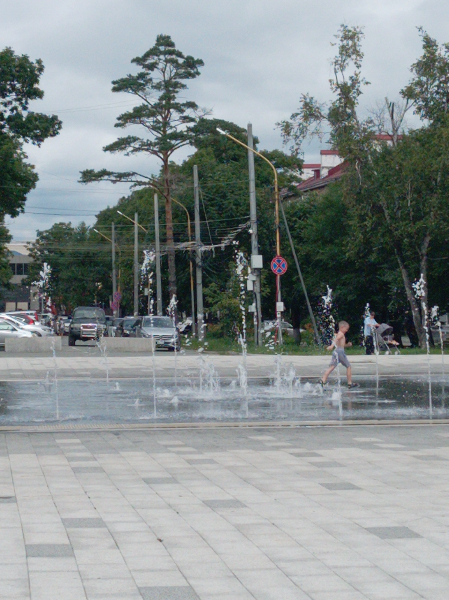 This is a full-frame, eye-level, outdoor, medium-shot photograph taken in Artyom, Russia. The scene depicts a public plaza with a functioning water fountain in the foreground. In the background, there are several cars parked on a street, and lush green trees and buildings line the street. A young boy, wearing shorts, is captured mid-stride in the fountain area, seemingly running through the water jets. Further in the background, a woman pushes a stroller. The sky is overcast, suggesting a cloudy day. The overall impression is a typical urban scene with recreational elements.