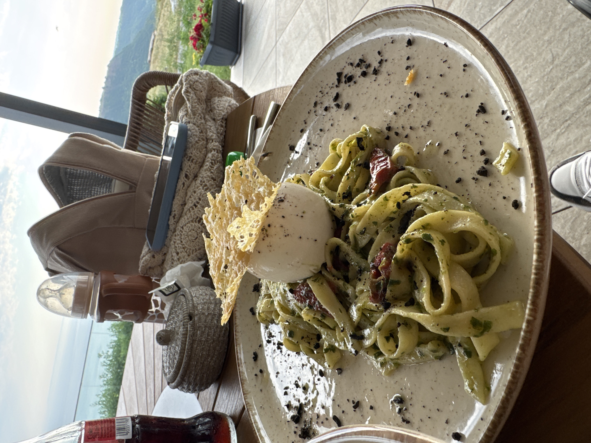 This media file captures a close-up of a plate of pasta served on a rustic wooden table. The pasta appears to be tagliatelle or fettuccine, tossed in a green sauce, possibly pesto, with visible chopped herbs and sun-dried tomatoes. A generous portion of burrata cheese sits atop the pasta, garnished with a delicate cheese crisp. The dish is sprinkled with what looks like black pepper or crushed olives, adding a touch of visual contrast.
In the background, blurred elements suggest an outdoor setting. A chair with a woven back and a knitted throw is visible to the left, along with a brown bag or backpack. Beyond this, a scenic view unfolds, with rolling hills and a hazy sky, indicative of a daytime scene, possibly late afternoon given the soft light. A glimpse of a balcony railing suggests the setting is elevated, perhaps a restaurant or cafe with a view.
To the left of the pasta, a baby bottle with a brown silicone holder is placed on the table, next to a small, woven decorative item. A partially visible bottle with a red label, possibly a drink, is in the foreground. The overall impression is a relaxed meal in a scenic location. The presence of the baby bottle hints at a family outing. No specific emotions are conveyed, but the scene suggests a moment of quiet enjoyment. The text on the bottle is too blurry to be legible. The location is identified as Kavarna, Bulgaria, which aligns with the hilly landscape.