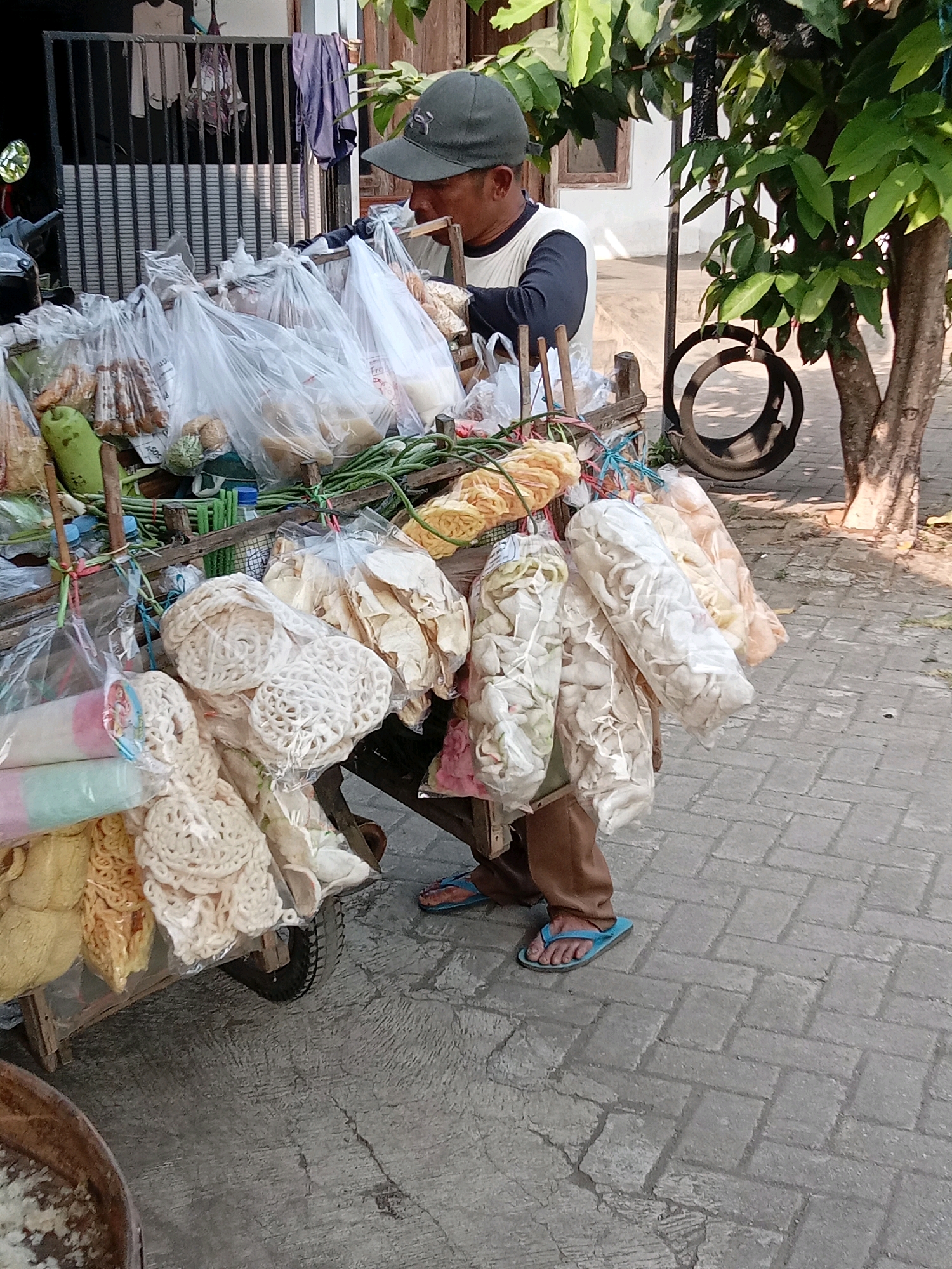The image depicts a street vendor in Sukoanyar, Indonesia, with a cart laden with packaged snacks and produce. A person, wearing a dark baseball cap, a long-sleeved shirt with white and dark sleeves, and brown trousers, is attending to the cart. They are seen reaching into one of the plastic bags containing the snacks. The cart itself is a wooden structure with a wheel, and it is filled with various items, including what appears to be puffed crackers, dried goods, and some fresh produce like long beans and a green gourd. The vendor is wearing blue flip-flops. The background shows a building with a window, some hanging clothes, and a tree with lush green leaves. The ground is paved with interlocking bricks. A large tire is hanging from a pole near the tree. The overall scene suggests a typical day for a street vendor in an Indonesian town.