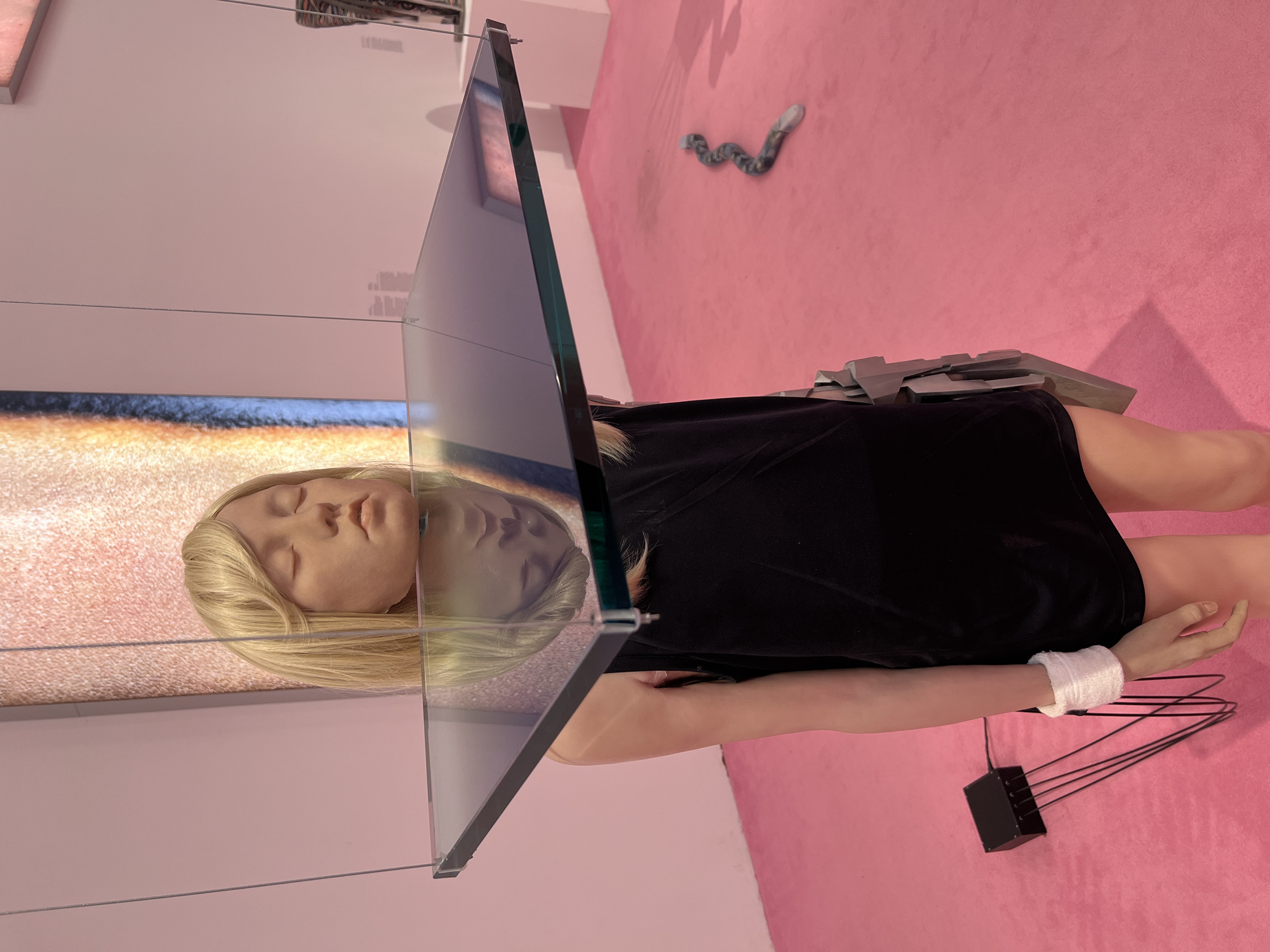 The image depicts an art installation within a gallery setting, likely in New York City. The primary subject is a life-size mannequin or sculpture of a blonde-haired individual, with eyes closed, positioned centrally. Above the head of this figure, a square pane of dark, reflective glass is suspended by thin wires, creating a mirrored surface that reflects the face of the mannequin. The mannequin is dressed in a black sleeveless garment and appears to be wearing a silver, angular prosthetic or accessory on its left hip. A white band is wrapped around the figure's left wrist, from which several black wires extend downwards to a small black electrical box situated on the pink carpeted floor. To the right and further back on the floor, a grey, serpentine object with colored segments lies coiled. The background consists of a pale pink wall, with a textured, vertical artwork or screen displaying warm tones of orange and yellow to the left of the central figure. The overall impression is one of contemporary art displayed in a clean, minimalist exhibition space.