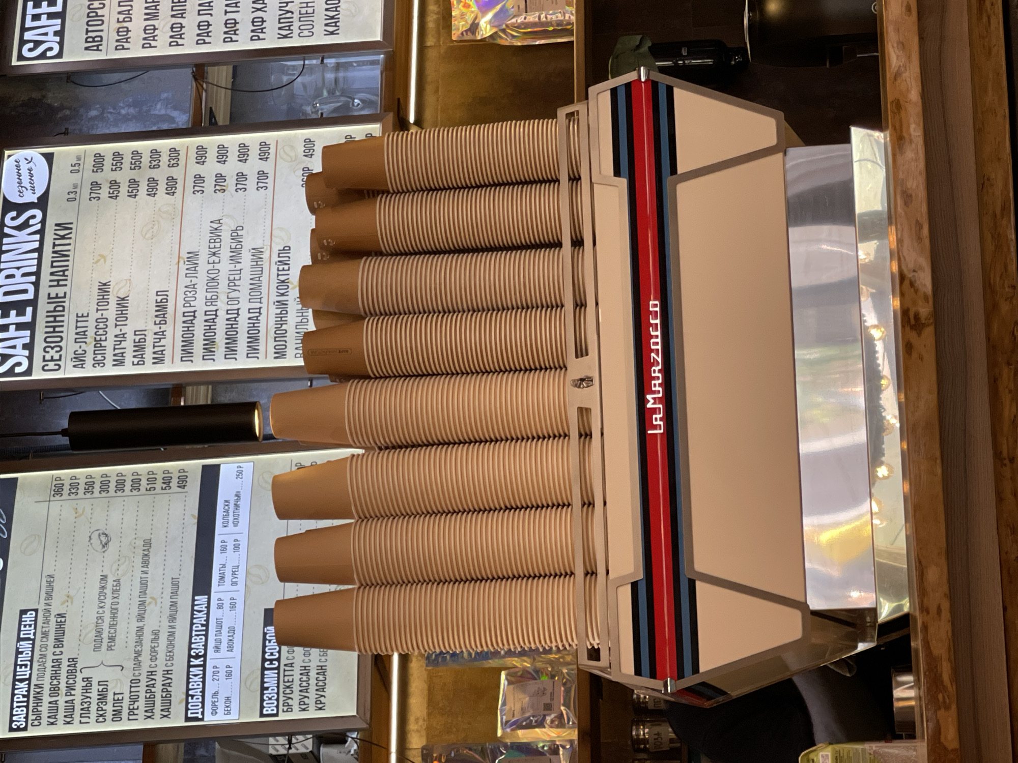 The image shows a close-up of a coffee machine with a stack of disposable cups in the foreground. The coffee machine is modern and has a distinctive design with vertical stripes of red, blue, and black on a beige background, along with the "LAMARZOCCO" logo. The cups are stacked in neat rows, suggesting a cafe or coffee shop setting.
To the left of the coffee machine, a menu board is visible, displaying a list of drinks and food items in Russian. The menu is illuminated and appears to be a digital display. It lists "SEZONNYE NAPITKI" (Seasonal Drinks), "ZAWTRAK CELIY DEN'" (Breakfast All Day), and "DOBAVKI K ZAVTRAKAM" (Additions to Breakfast), along with prices in rubles. Some of the items listed include "AYC-LATTE," "ESPRESSO-TONIK," "MATCHA-BAMBL," "LIMONAD ROZA-LAYM" (Rose-Lime Lemonade), "SIRNIKI," "KAWA OVSYANAYA" (Oatmeal Porridge), and "KRUASSAN" (Croissant). The presence of food items and drinks suggests this is a cafe.
The background is dark and somewhat out of focus, with some lights and reflections visible. The overall impression is that of a busy, modern cafe. The image is oriented vertically. The setting is a cafe in Zelenogradsk, Russia.