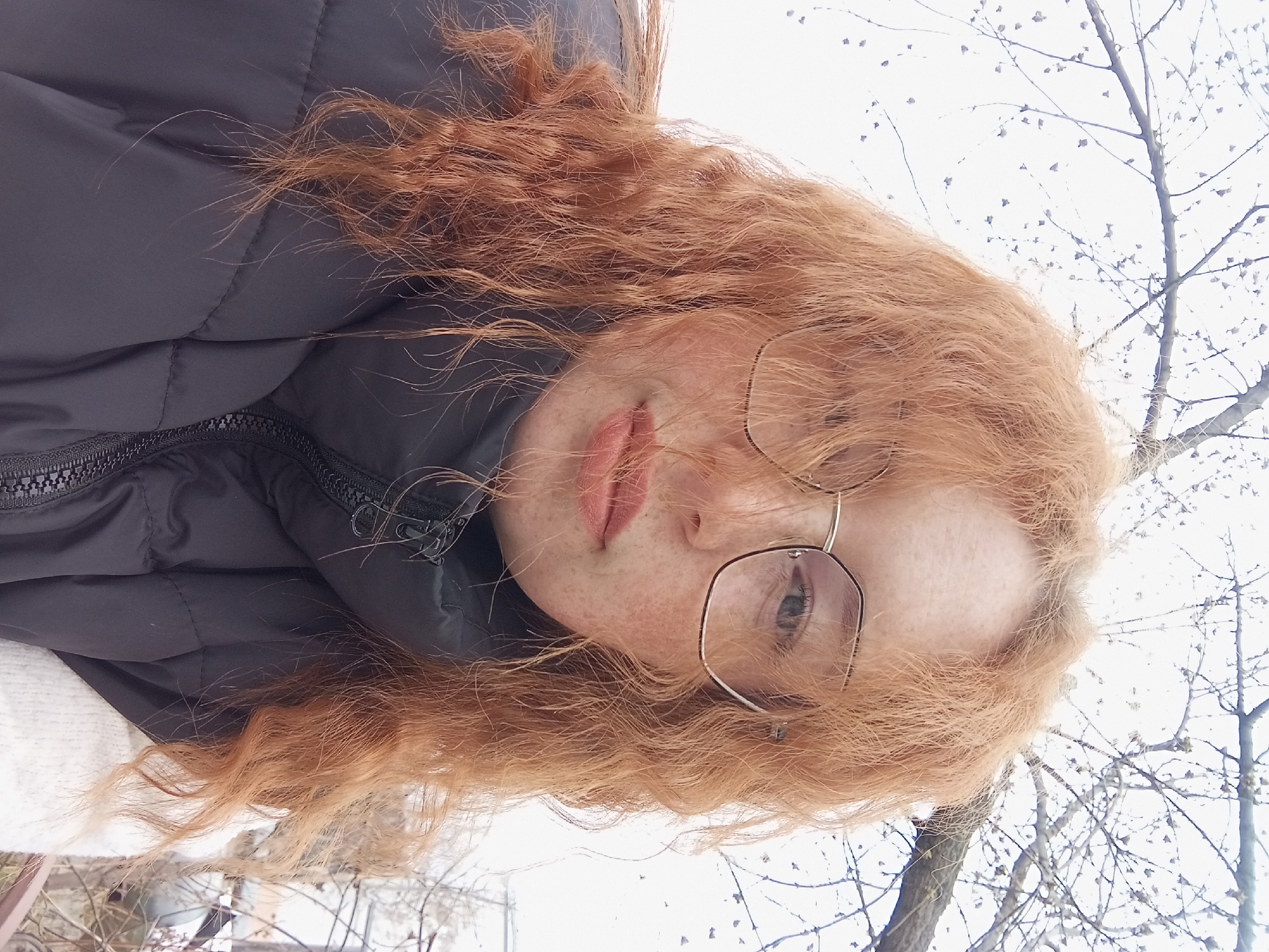 A person with red, curly hair and freckles is wearing glasses and a black puffer jacket. The background appears to be outdoors during winter in a city, with bare trees visible against a light sky. Location unknown in Yurgamysh, Russia.