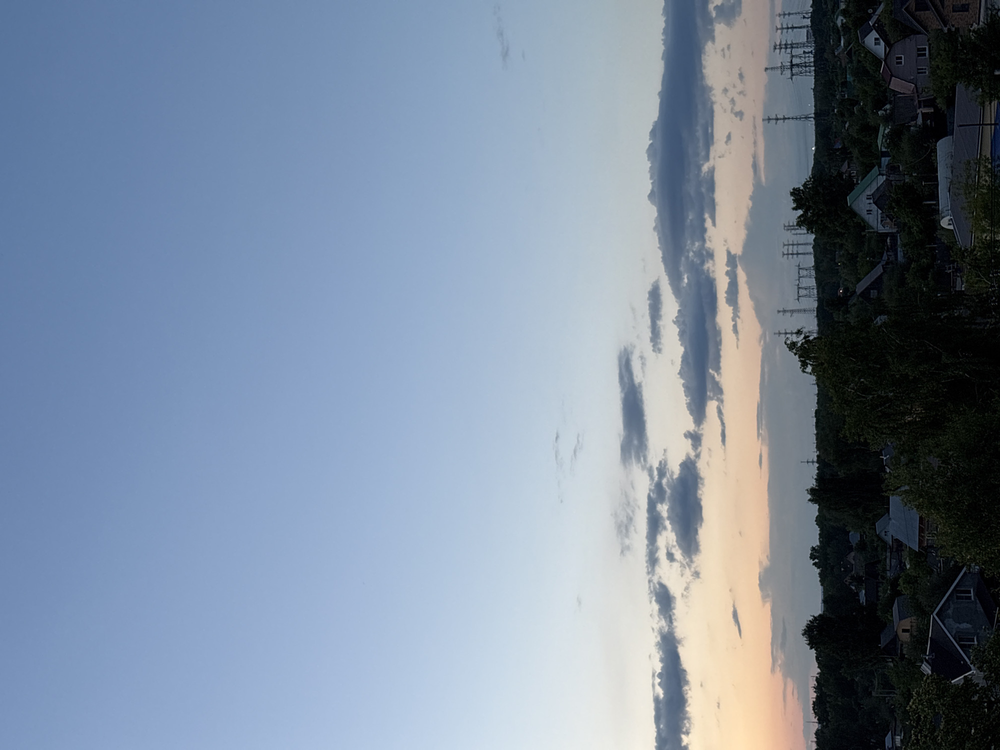 The image captures a panoramic view of a residential area in Chaikovsky, Russia, under a vast, evolving sky. The scene depicts a tranquil landscape at either sunset or sunrise, suggested by the soft, transitioning colors of the atmosphere.
The sky occupies the majority of the frame, moving from a clear, light blue in the upper left to a warm, soft peach and orange hue along the horizon on the right side. Scattered across the sky are several elongated, dark grey clouds, particularly concentrated towards the horizon, some of which are illuminated from below by the warm light.
Below the expansive sky, the horizon features a dark band of dense green trees, interspersed with residential houses. These houses exhibit various roof colors, including dark grey, green, and brown, indicating a suburban or rural setting. Further in the distance, a body of water, possibly a river or lake, stretches across the landscape, reflecting the sky's light. Prominently visible above this distant area are numerous tall, lattice-like structures, identified as electricity transmission towers or power line pylons, silhouetted against the lighter parts of the horizon.
No specific activity or event is taking place, and there are no people visible. The weather appears calm and partly cloudy. There is no visible text in the image. The overall mood conveyed is one of peace and serenity as daylight fades or begins.
