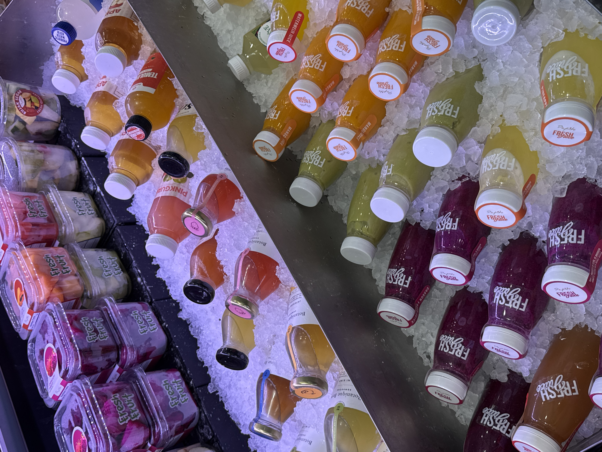 The image shows a refrigerated display case filled with assorted bottled juices and small plastic containers of cut fruit, likely in a retail setting such as a grocery store or convenience store. The juices are in various colors and are submerged in crushed ice. Some bottles have white caps, while others have black or gold caps. Labels on the bottles indicate flavors like "Mango Juice", "Watermelon Juice", and "Dragon Fruit Juice". The fruit containers are stacked and appear to contain colorful chopped fruits, with labels indicating "Fresh Fruit". The overall environment is clean and well-lit, with a focus on the chilled products. The scene is devoid of people. The location is identified as North Kuta, Indonesia, suggesting a tropical climate where chilled beverages and fresh fruit are popular.