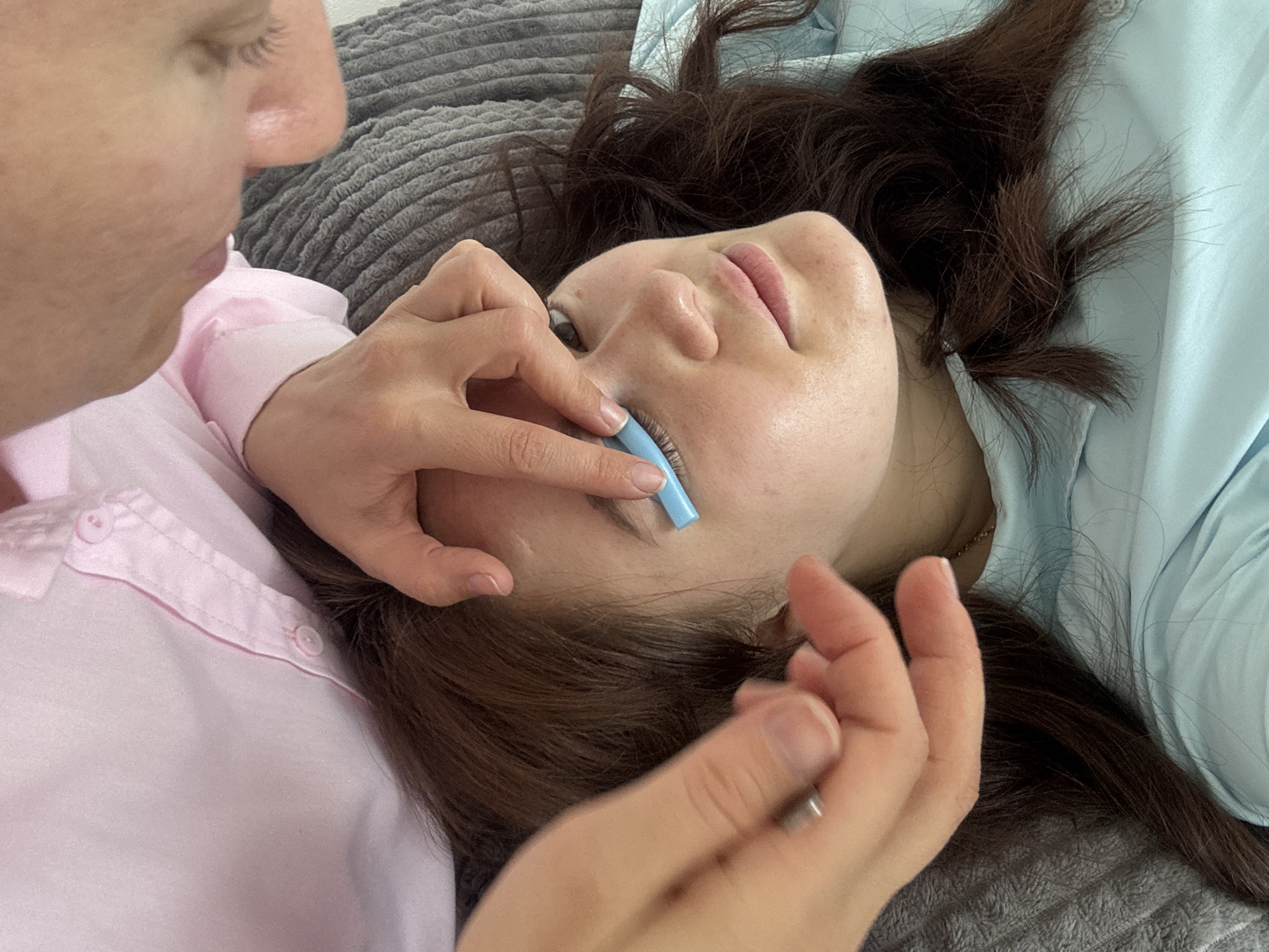 A close-up, high-angle shot shows a young woman lying down, her head on a gray textured surface. She has long, dark brown hair and is wearing a light blue shirt. A person wearing a pink collared shirt is leaning over her, with their hands performing a procedure on the woman's left eye. One hand, with a silver instrument, is holding the lower eyelid down, while the other hand is using a light blue, curved object to lift and separate the eyelashes. The scene appears to be indoors, possibly in a beauty salon. There is no information in the image to determine the specific location in the city of Chaikovsky, Russia.