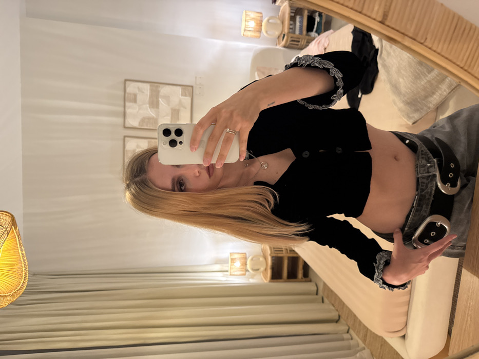 A woman stands in front of a mirror, using her phone to capture a selfie. She's wearing a black crop top with ruffled sleeves, denim jeans, and a double-buckle belt. The background shows a bedroom setting, featuring a bed, curtains, and decorative wall art. A rattan mirror frame adds texture to the composition. The overall lighting suggests a warm, indoor atmosphere.
