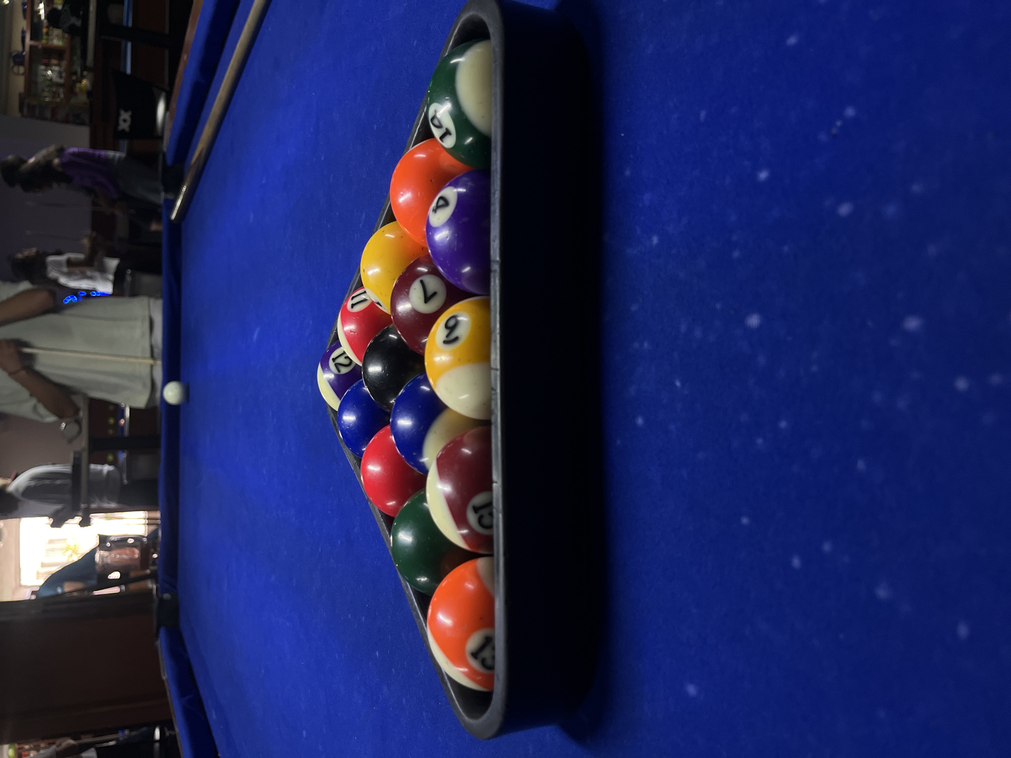 The image captures a pool table with a blue felt surface, arranged for a game of billiards. A black triangular rack holds numerous numbered pool balls tightly together, prominently displaying various solids and stripes. Visible numbers on the balls include 15, 8, 7, 9, 12, 13, 1, 10, 3, 11, and 14. A white cue ball is positioned near the left edge of the table, and a pool cue rests along the table's side.
In the blurred background, several people are visible, suggesting an active social setting. One person in a purple top is leaning over, seemingly engaged in the game, while another in a white top appears to be holding a pool cue. The setting appears to be an indoor recreational area, likely a bar or pub, with dark furniture and shelves holding bottles visible in the dimly lit, out-of-focus background. The image is oriented vertically, giving a dynamic perspective of the setup. The blue felt of the table shows some minor dust or wear.