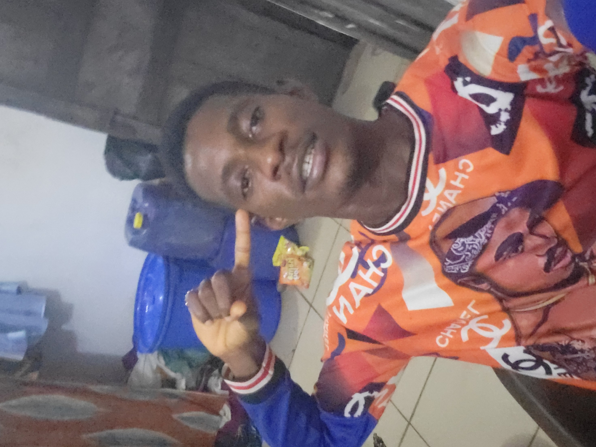 The image shows a young Black male posing for a selfie in an indoor setting. He is wearing an orange long-sleeved shirt with a repeating pattern of the Chanel logo and images of Tupac Shakur. The background appears to be a room with some large blue plastic containers, a striped curtain, and what looks like clothes hanging in the upper left corner. The man is holding his left hand up to his temple. He is the primary subject of the photograph.