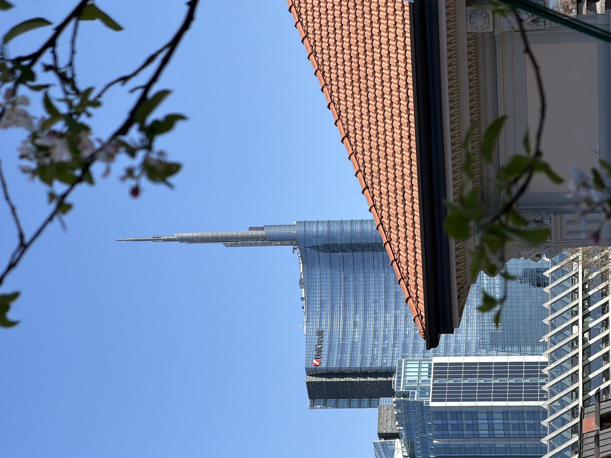 The image captures a view of the UniCredit Tower in Milan, Italy, framed by architectural elements including a terracotta tiled roof and decorative moldings. The tower, a modern glass skyscraper with a distinctive spire, stands tall against a clear blue sky. Branches with blossoms in the foreground and background provide a sense of depth and seasonal context. Additional buildings with glass facades are visible, partially obscured by the main structures in focus.