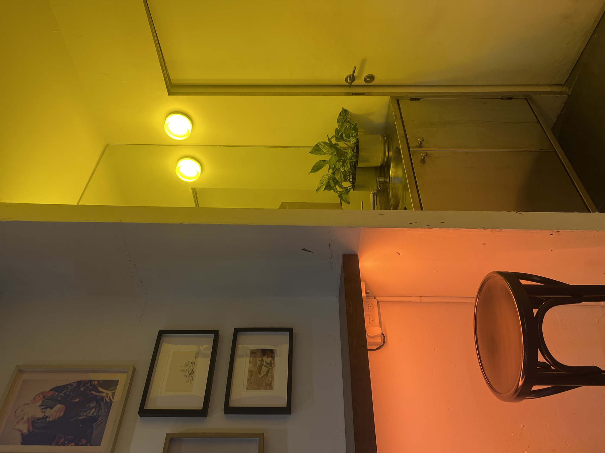 An interior scene of a possible kitchen in Tel-Aviv, Israel, is presented in an upside-down perspective. The scene includes a counter with a sink, cabinets, a plant in a pot, two recessed lights, and a door. On the lower half of the upside-down perspective, there are three framed pictures on a wall, an electrical outlet, and part of a wooden chair.