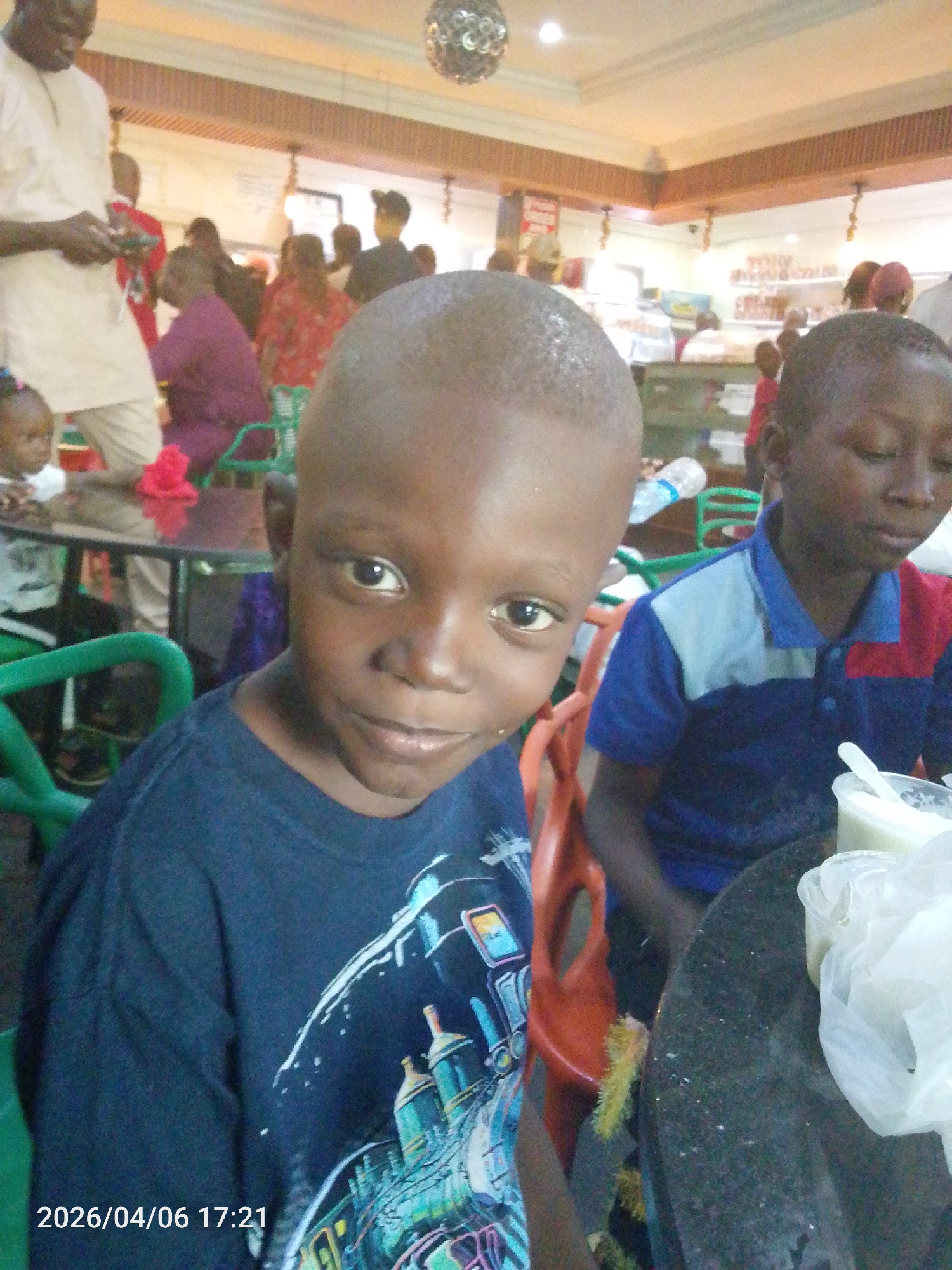 The image shows two young boys seated at a table in what appears to be a restaurant or cafeteria. The boy in the foreground is wearing a dark blue T-shirt and is looking directly at the camera. The boy in the background is wearing a blue and red polo shirt, and is looking down at something on the table. On the table, there are food containers and a spoon. Other people are visible in the background, along with tables, chairs, and what looks like a food display case. The environment appears to be indoors, and the timestamp on the image indicates a date in the future. There is no information in the image that indicates that the location is in Lokoja, Nigeria.