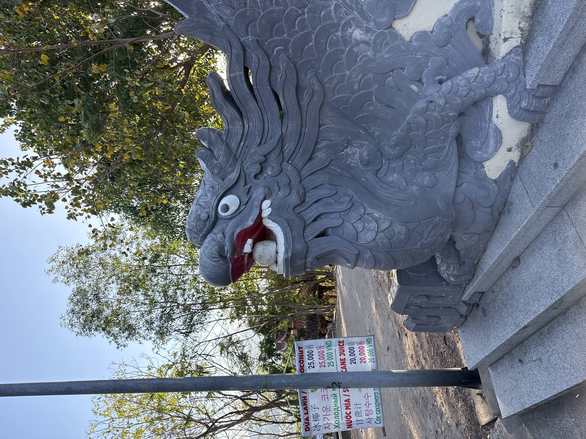 The image shows a close-up of a dragon sculpture, possibly part of a gate or entrance feature, in what appears to be the Nha Trang Ward area of Vietnam. The sculpture is gray and detailed, with prominent scales and a somewhat comical expression. Below the sculpture is a sign listing prices for "OCONUT" and "CANE JUICE" in Vietnamese, Chinese, Korean, and Russian, with the prices indicated in VND. The sign is tied to a metal pole. Background foliage suggests a park or public space.