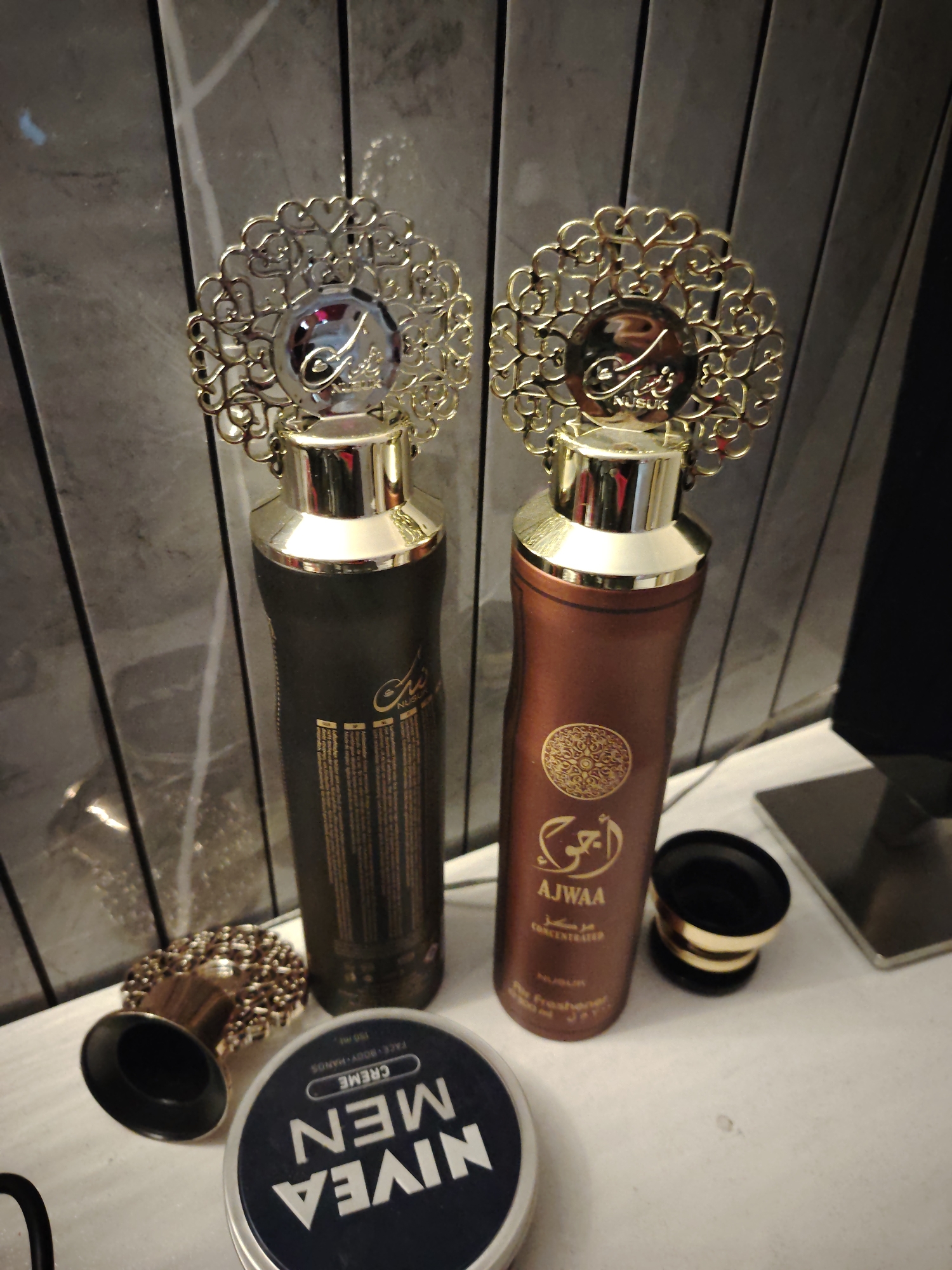 This image depicts a collection of cosmetic and fragrance products displayed on a white surface against a dark grey paneled background, likely situated indoors in the city of Huntingdonshire, United Kingdom.
In the center of the frame, two tall, slender bottles stand upright. The bottle on the left is dark brown or black with gold accents, featuring a gold cap and an elaborate, circular gold filigree decoration at its top. This decoration centers on a reflective gold medallion bearing intricate script, with "NUSUK" clearly visible underneath. The bottle's body displays gold Arabic script and fine-print English text, with "NUSUK" also discernible. The bottle on the right is a reddish-brown or bronze color, identical in form and adornment to the left bottle, including the gold cap, ornate filigree, and "NUSUK" medallion. Its body clearly shows prominent gold Arabic script, followed by "AJWAA" and "CONCENTRATED", and further down "NUSUK Air Freshener 300 ml". A decorative, circular gold pattern is also visible on its upper body.
To the left of the brown bottle, on the white surface, rests a dark blue circular tin lid of "NIVEA MEN CREME" with "FACE BODY HANDS 150 ML" printed on it. Adjacent to this, to the right of the Nivea tin, is the inverted base of a small, decorative container, featuring a similar gold filigree pattern around its edge. The matching black and gold lid of this container stands upright behind the brown "AJWAA" bottle.
The items are arranged on a clean, light-colored, possibly white or off-white, reflective surface. The background consists of vertically oriented, dark grey panels, possibly a wall, with subtle lighter grey or white striations, suggesting a stone or textured material. No individuals are present, and no actions are being performed.