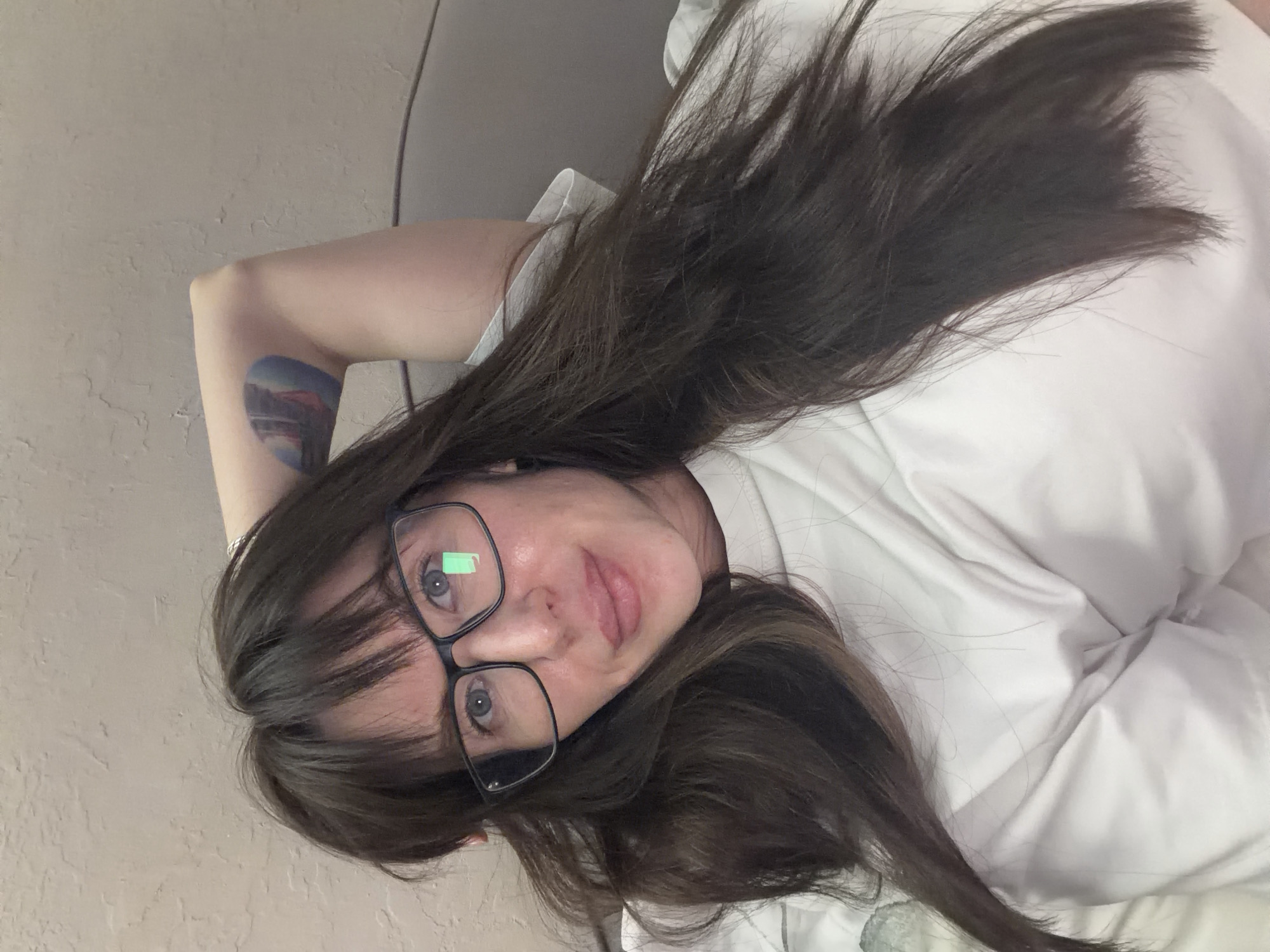 A woman with dark hair and glasses is lying down, presumably indoors, in a white shirt. She has a tattoo on her left arm. The location is unknown, and there's no indication of Perm, Russia.