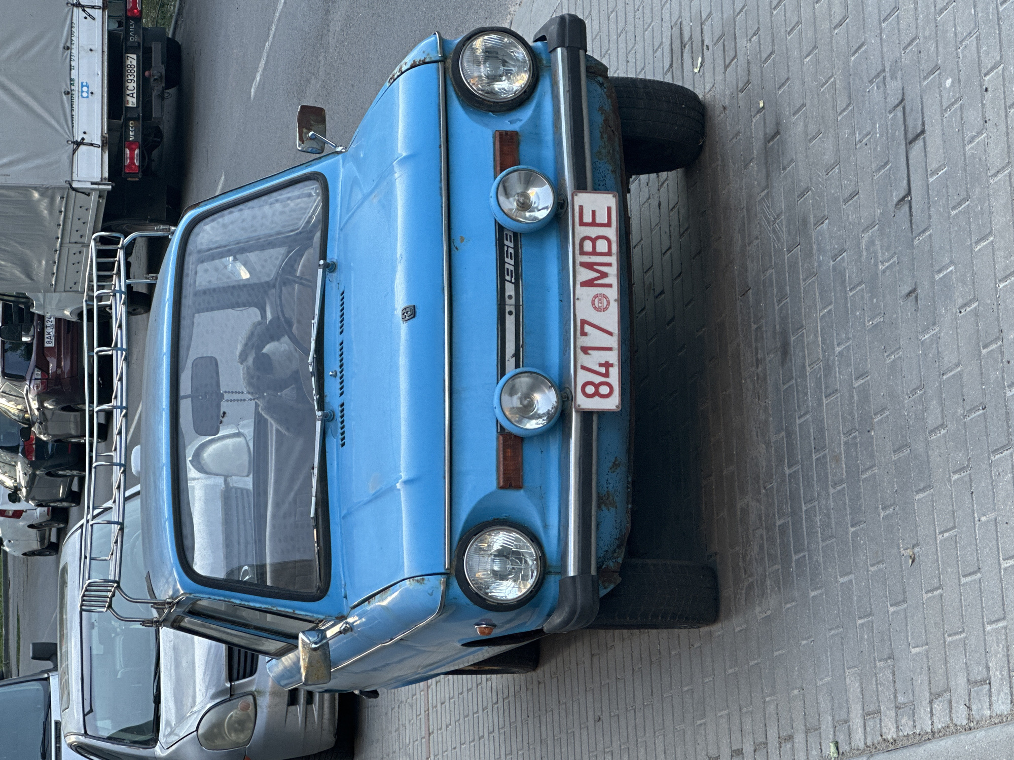 The image captures a light blue vintage car, likely a 1968 model, parked on a brick-paved surface. The car features round headlights, a vertical chrome grille, and a license plate that reads "8417 MBE BELARUS." The background includes other parked vehicles, including a white truck with "AC 9388-7" on its license plate. The location appears to be a street in Minsk, Belarus, based on the license plate. The photo is taken at an angle, with the car slightly rotated.