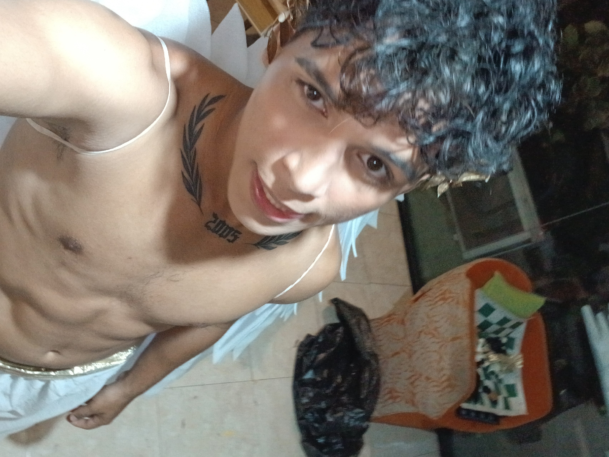 A medium-shot, high-angle selfie shows a shirtless young man with short, curly black hair and brown eyes, posing with a slight smile. He appears to be wearing a costume, possibly as an angel, evidenced by the white straps and what seem to be white wings partially visible behind him. He has a tattoo of leaves on his upper chest and the number "2005" inked on his neck. A gold-colored belt is worn low on his waist. The background is a slightly cluttered interior space with a tiled floor, a dark plastic bag, and what looks like a storage bin or chair with a patterned cloth on it. It is dark. The location is Playa El Agua, Venezuela.