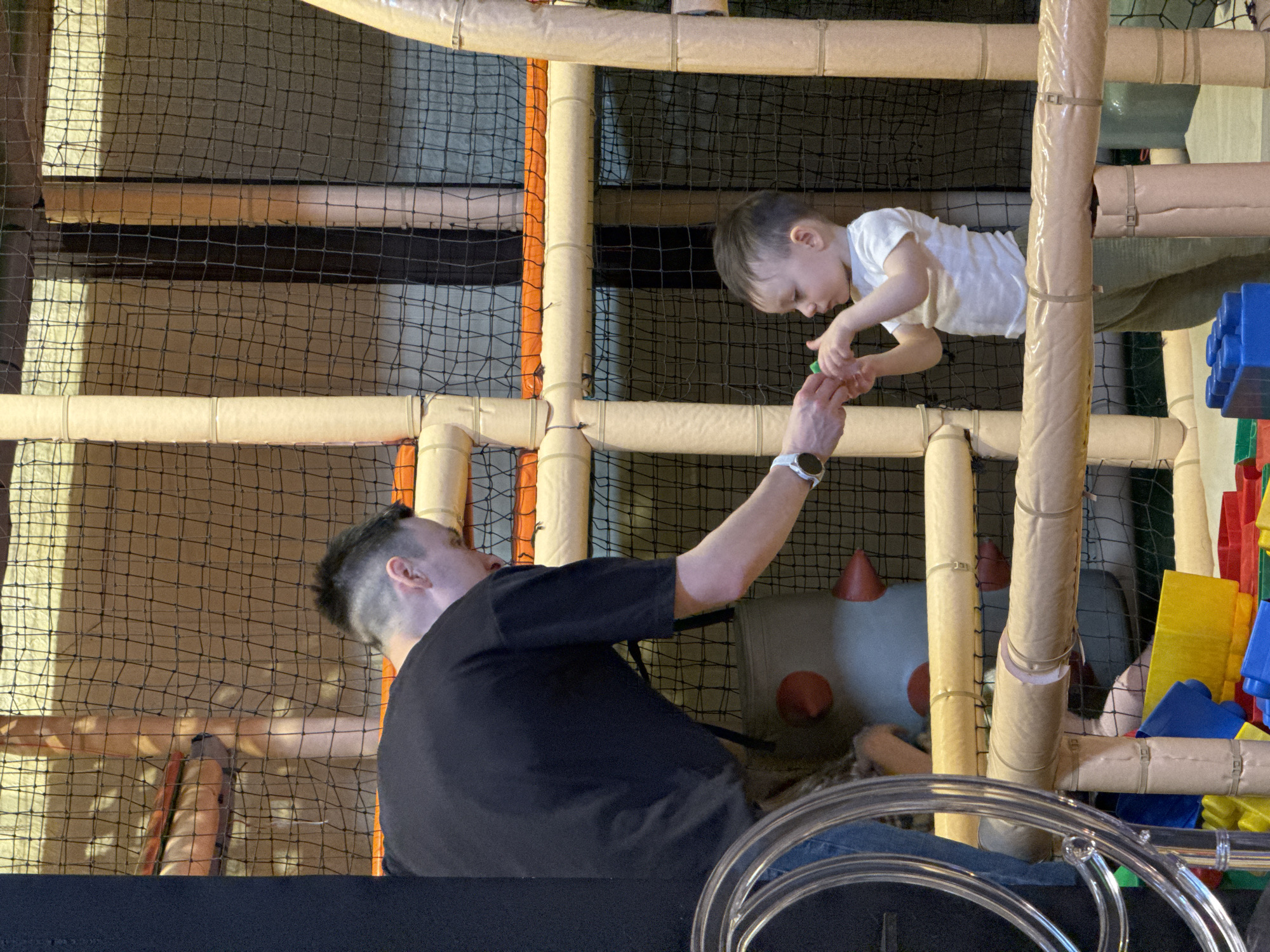 The image depicts a man and a child interacting within an indoor playground setting. The man, wearing a black t-shirt and jeans, is extending his arm upwards towards the child. The child, dressed in a white t-shirt and grey pants, appears to be reaching down to receive something from the man. The playground structure is constructed of netting, padded pipes, and colorful plastic blocks. The scene is in the city of Izhevsk, Russia.