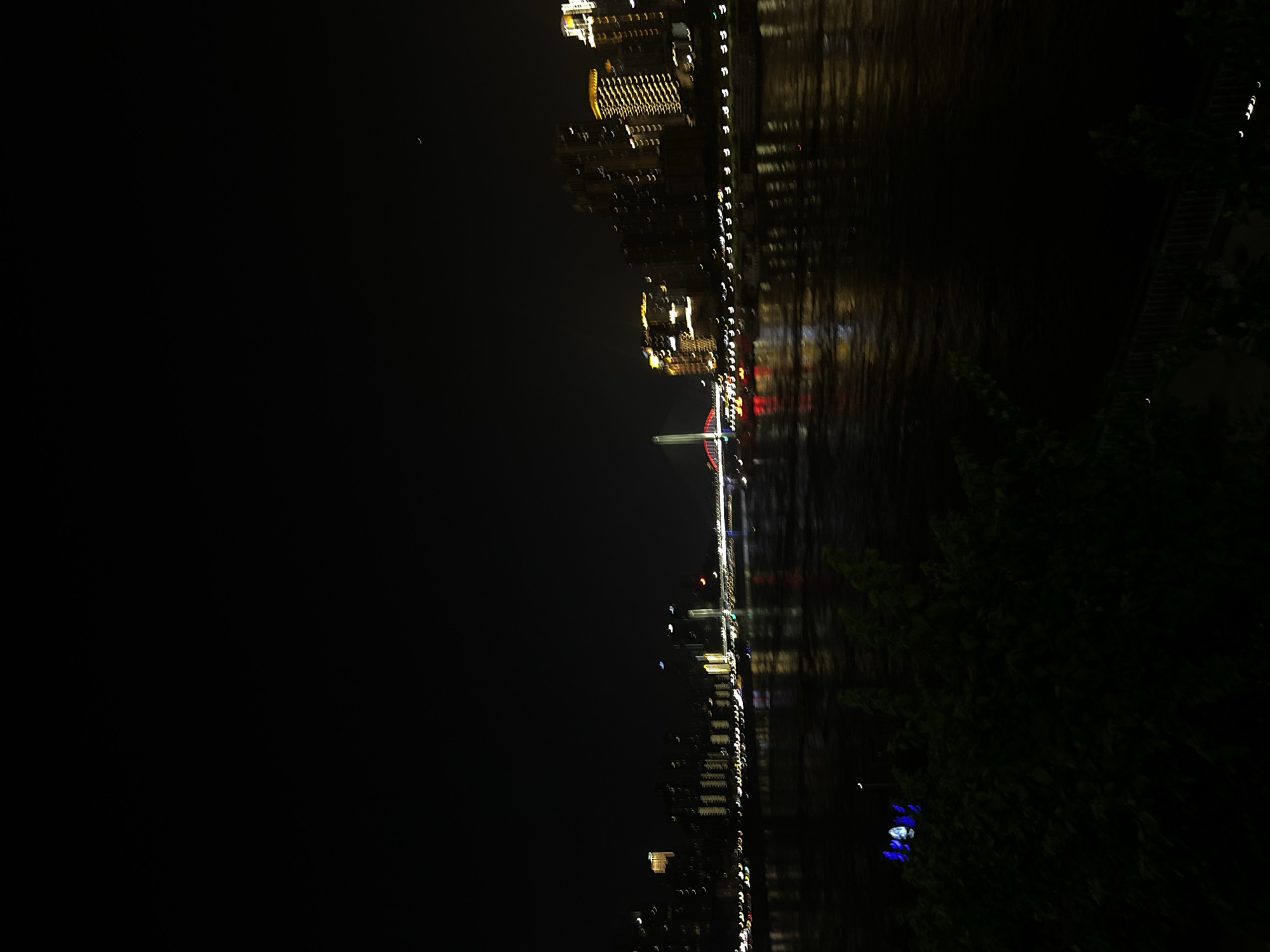 This is a vertically oriented nighttime photograph taken from a slightly elevated perspective, looking down at a waterfront scene. The image captures the illuminated cityscape reflected in dark, calm water.
On the left side of the frame, a line of buildings is visible, characterized by numerous windows lit with warm, yellow light. These buildings appear to be tall and modern, with their reflections stretching into the water below. Some of the building lights are blurred due to motion or the exposure of the photograph, creating a painterly effect.
The water takes up the right two-thirds of the image and shows distorted reflections of the cityscape. The dark surface of the water is broken by streaks of light, indicating ripples or movement. In the distance, a red-lit structure, possibly a bridge or a tower, stands out against the night sky.
On the far right, a dense mass of dark green foliage, likely trees or bushes, occupies the lower portion of the frame. Their dark forms contrast with the illuminated cityscape. A few small, blue lights are visible within the foliage.
The overall impression is one of a vibrant city at night, with the lights creating a dazzling spectacle against the dark sky and water. The time of day is clearly night, and the presence of city lights and reflections suggests an urban setting. The calm water implies still weather conditions. There is no visible text in the image. The scene evokes a sense of nighttime beauty and urban energy.