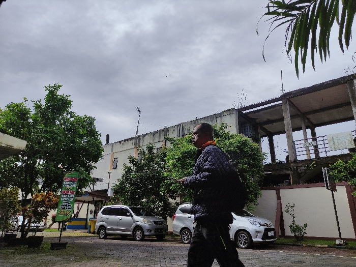 The image depicts an outdoor urban scene in Kepanjen, Indonesia, under an overcast sky. A man walks from left to right across a paved area in the foreground. He is seen in profile, wearing a dark, possibly quilted, jacket, dark trousers, and carrying a dark backpack. An orange collar or scarf is visible around his neck. His expression is neutral as he looks forward.

To the left, a green upright sign with white and yellow text is prominent. Readable text includes "MINGGU RAMAI" (Crowded Sunday), "HARGA..." (Price...), "TERSEDIA" (Available), "MINUMAN" (Drinks), "JUGA" (Also), "ROKOK" (Cigarettes), "DLL" (etc.), "CUCI SALJU" (Snow Wash - a car wash method), "MELAYANI" (Serving), "JUAL PULSA" (Selling credit/top-up), "KUOTA" (Internet data), and "GAME." A graphic of a car is also visible on the sign.

Behind the sign and man, a silver multi-purpose vehicle (MPV) and a silver hatchback are parked on the paved surface. Lush green trees are scattered throughout the scene, notably on the left, right, and in the mid-ground behind the cars. In the background, there are two buildings: a light-colored multi-story building on the left, and on the right, an unfinished or dilapidated concrete structure with exposed pillars and slabs, suggesting multiple levels. A low wall separates the foreground from this structure. Palm fronds hang into the frame from the upper right corner. The sky is entirely covered with gray clouds, indicating a cloudy daytime.