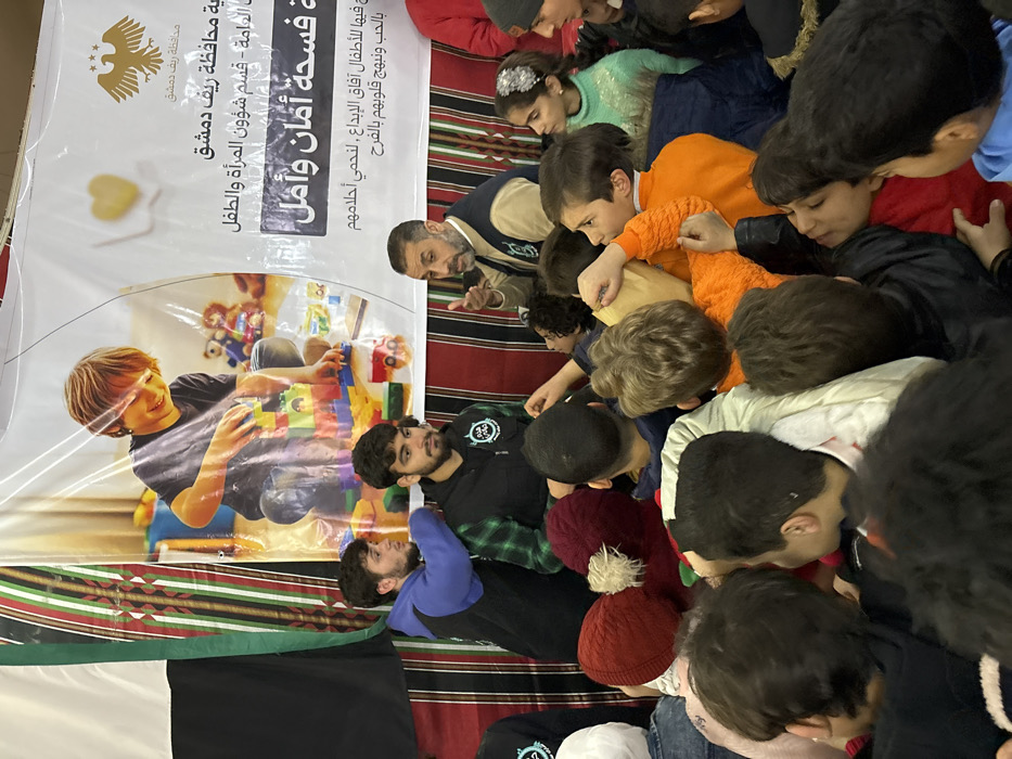 This image captures an indoor community event in Kafr Batna, Syria, featuring a large group of children interacting with several adults. In the background, a prominent white banner displays an image of a smiling child playing with colorful building blocks. Arabic text on the banner identifies it with the "Damascus Countryside Governorate," "Women and Child Affairs Department," and states "A Space of Safety and Hope," accompanied by an eagle emblem. Behind and to the sides of the banner, a patterned fabric, resembling traditional textiles in shades of brown, black, white, and green, is visible, alongside a partial view of the Syrian flag on the far left.
In the mid-ground, an adult male with a beard, wearing a light brown vest over a patterned shirt, is actively speaking and gesturing with his right hand towards the assembled children. To his immediate left stand two younger adult males; one with a beard, wearing a dark vest over a green plaid shirt with a logo, and another in a blue hoodie over a dark top, looking towards the speaking adult. They appear to be staff or volunteers.
The foreground and mid-ground are densely populated by approximately two dozen children, predominantly young boys and some girls, all facing towards the adults. They are dressed in various casual winter clothing, including jackets, sweaters, and knitted hats, such as a child with a red hat and a white pom-pom. Several children are attentive to the speaking adult, with some raising their hands or reaching forward, indicating an interactive or distribution event. A girl on the far right, wearing a light green sweater and a sparkling hair accessory, observes the scene. The overall scene depicts an organized gathering for children in what appears to be a community setting.