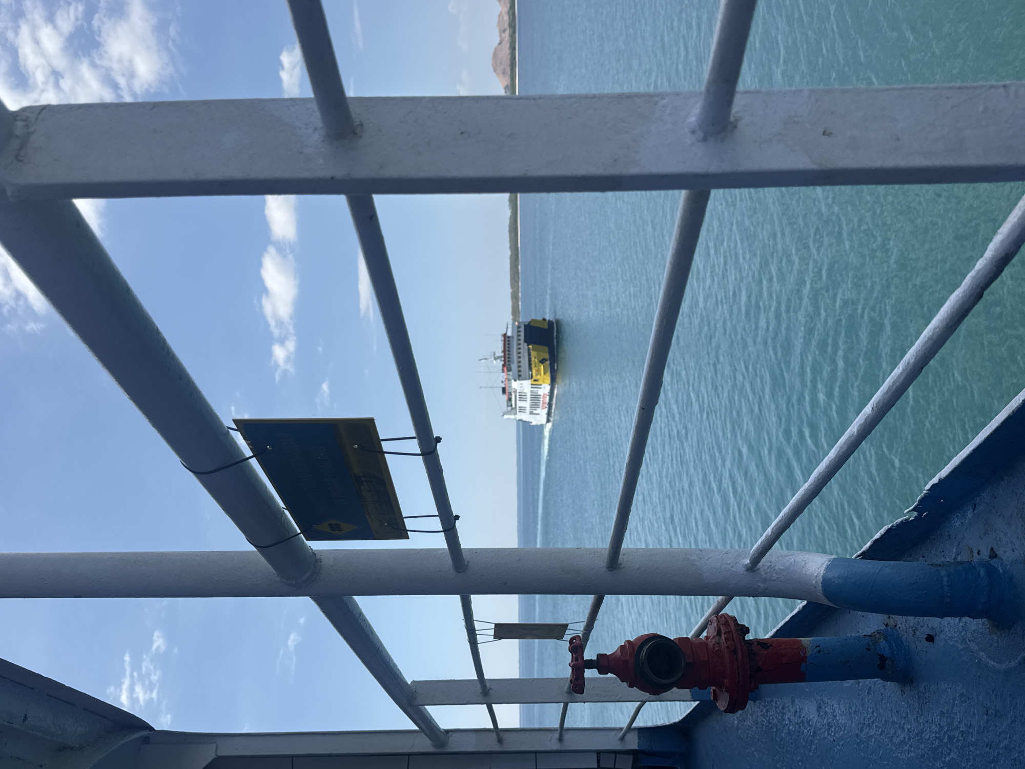 The image depicts a view from a ship's deck, looking out at a calm, turquoise sea under a bright blue sky with scattered white clouds. In the distance, a white and yellow ferry is visible, moving away from the viewer. The foreground shows the blue deck of the ship, with a red fire hydrant attached to a post. Metal railings, painted white, divide the view of the sea. A yellow and black sign with text in a foreign language is attached to one of the railings. In the far background, a light brown, hilly coastline can be seen. The scene is set in the waters near Upt Tambak Sari, Indonesia.