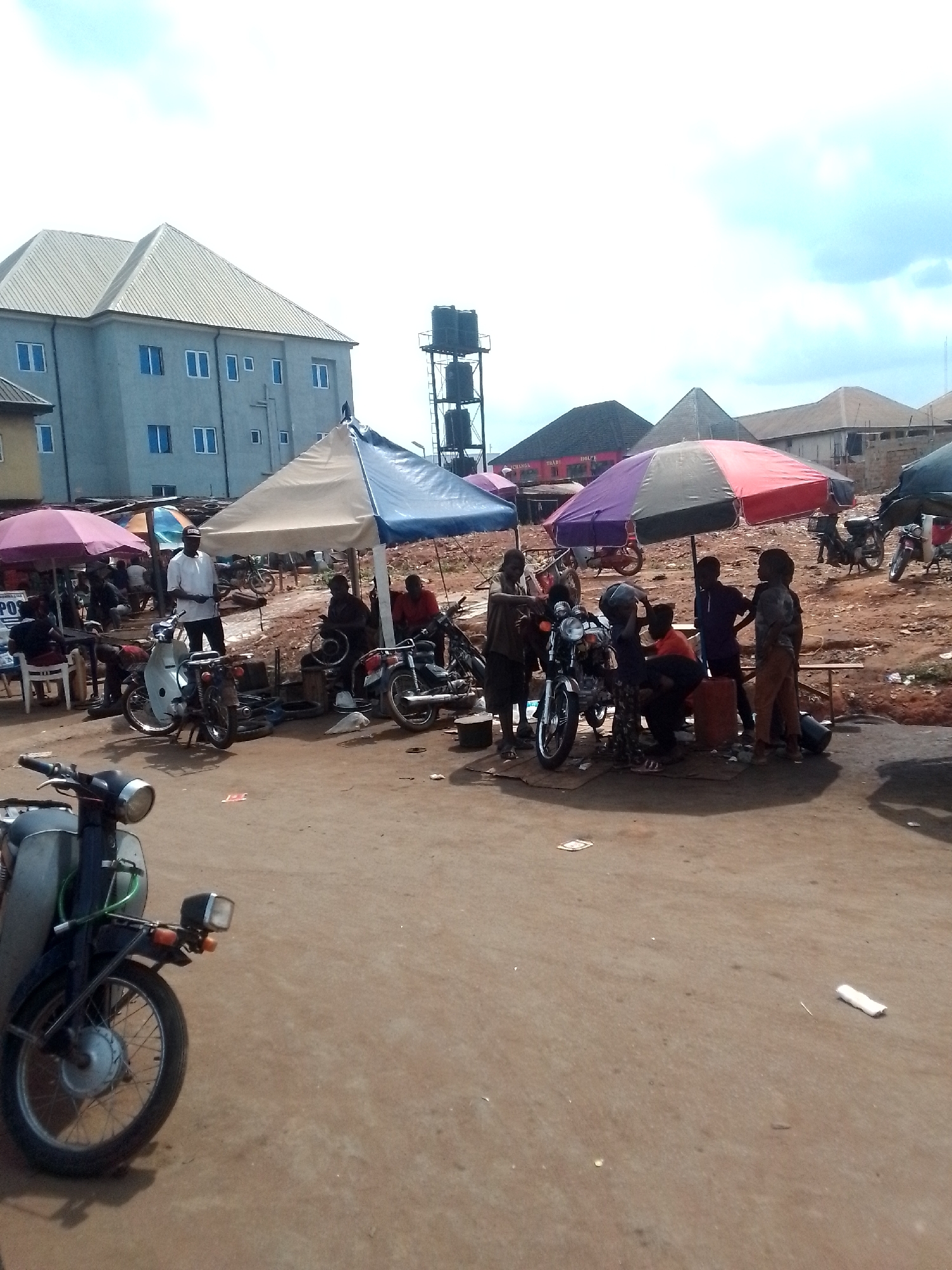 This image depicts a bustling outdoor scene, likely a marketplace or a roadside repair area in Orlu, Nigeria. A group of people are gathered around several motorcycles, some under makeshift shelters provided by large umbrellas. Some individuals appear to be working on the motorcycles, possibly repairing or maintaining them, while others are observing. The setting is an open space with a dirt ground, typical of many urban areas in Nigeria. In the background, there are buildings and water storage tanks, which offer context to the location. A man in a white shirt stands near one of the motorcycles. Several small children are also present, watching the activities. The overall impression is of a busy, informal economic activity taking place in a public space.